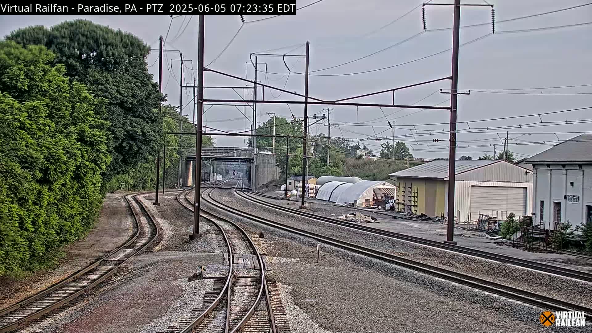 Multiple sets of train tracks curve through a landscape of green trees, industrial buildings, and overcast skies.