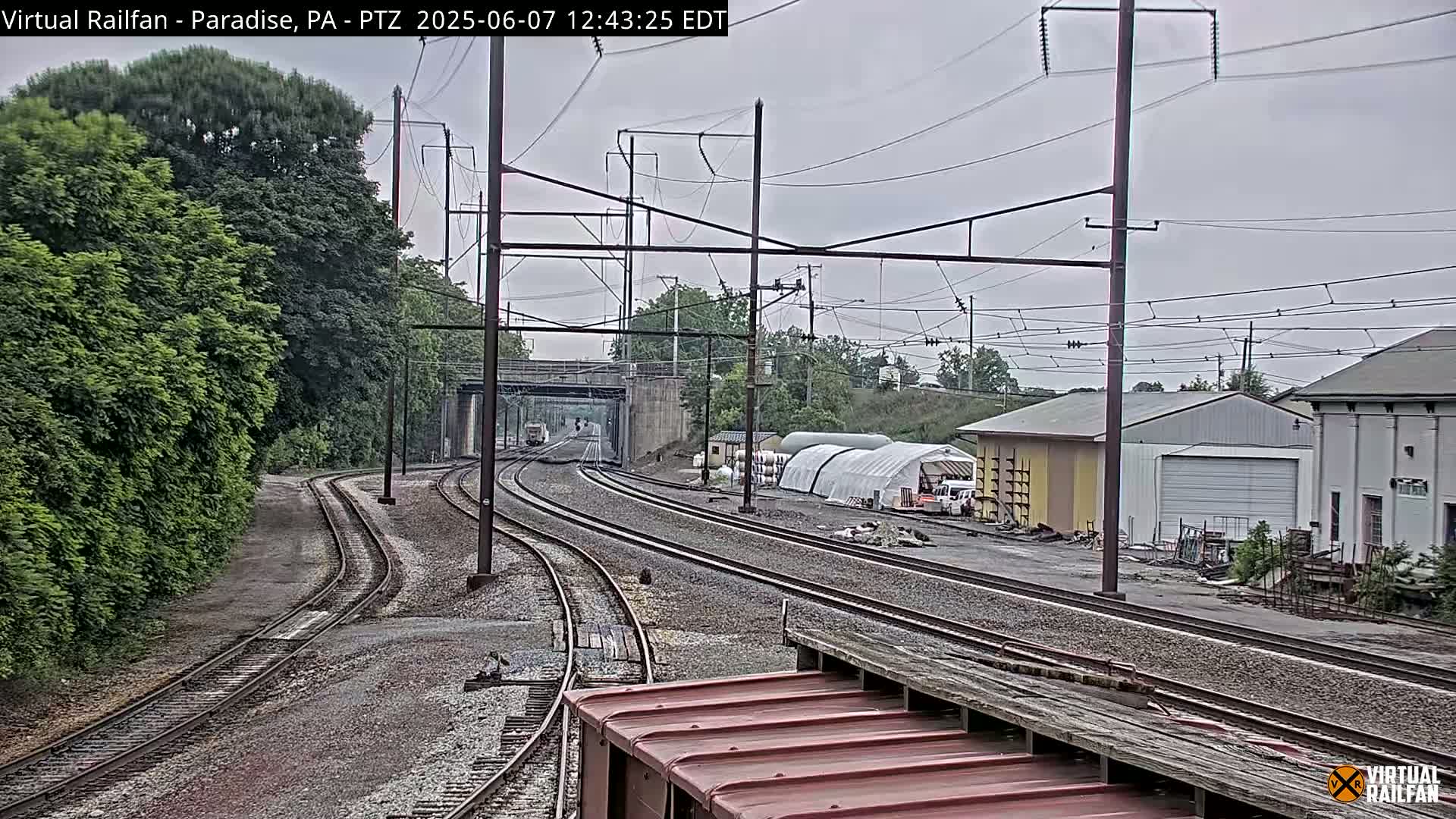 Multiple train tracks extend into the distance under an overcast sky, passing by a few buildings and lush green trees.
