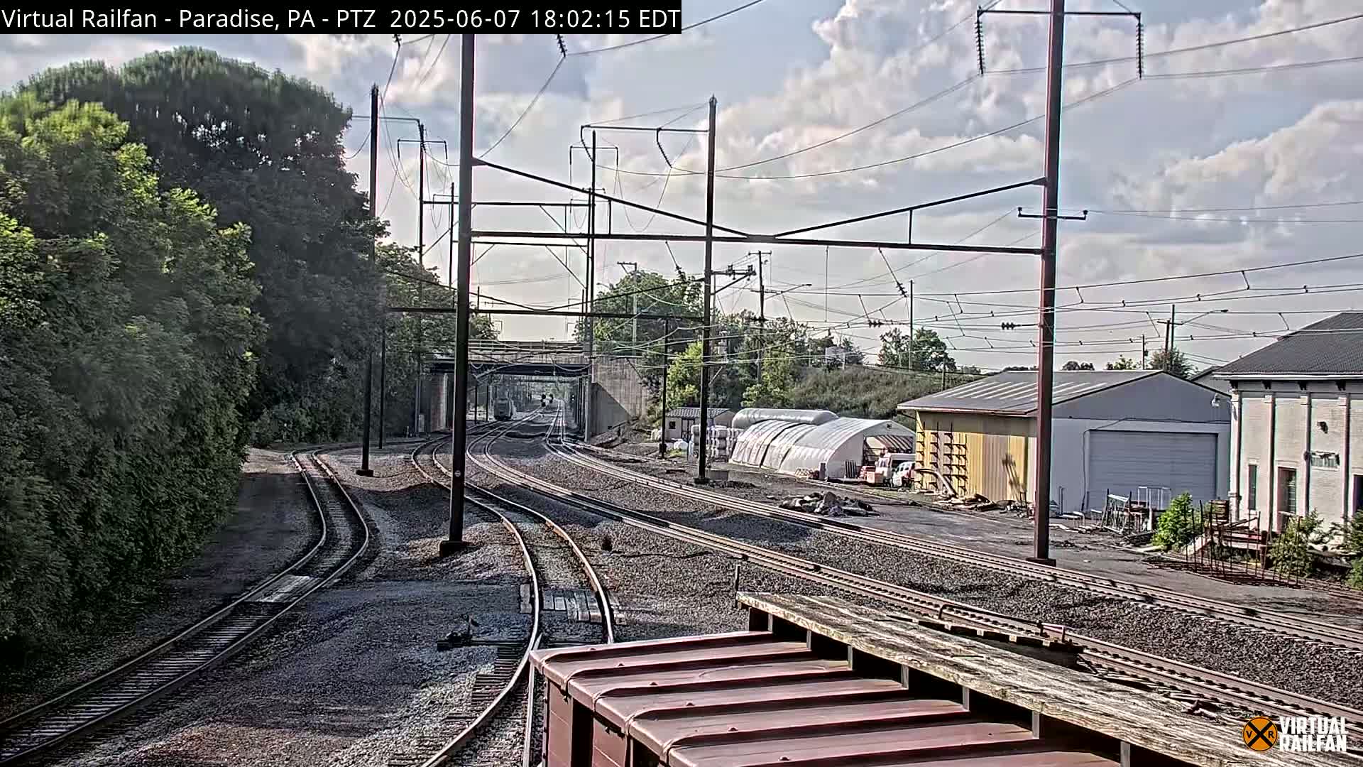 Multiple train tracks extend into the distance under a partly cloudy sky, passing alongside trees, buildings, and a large shed.