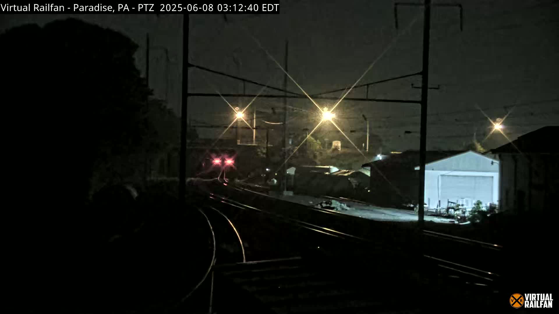 A nighttime view of railroad tracks with a train in the distance, under lightly falling rain or snow, illuminated by several streetlights.