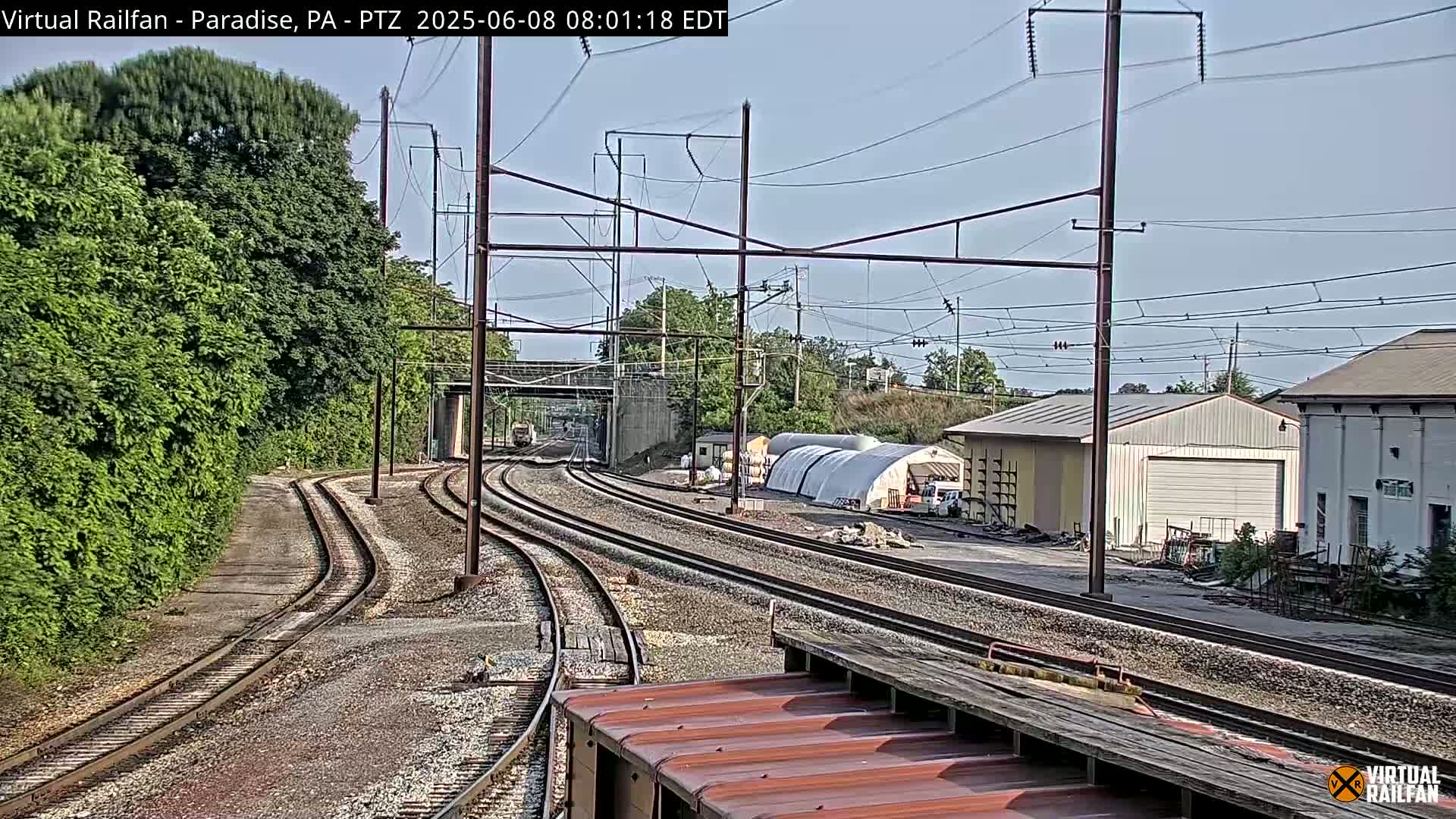 Several train tracks curve through a landscape of green foliage, passing industrial buildings under a mostly clear sky.
