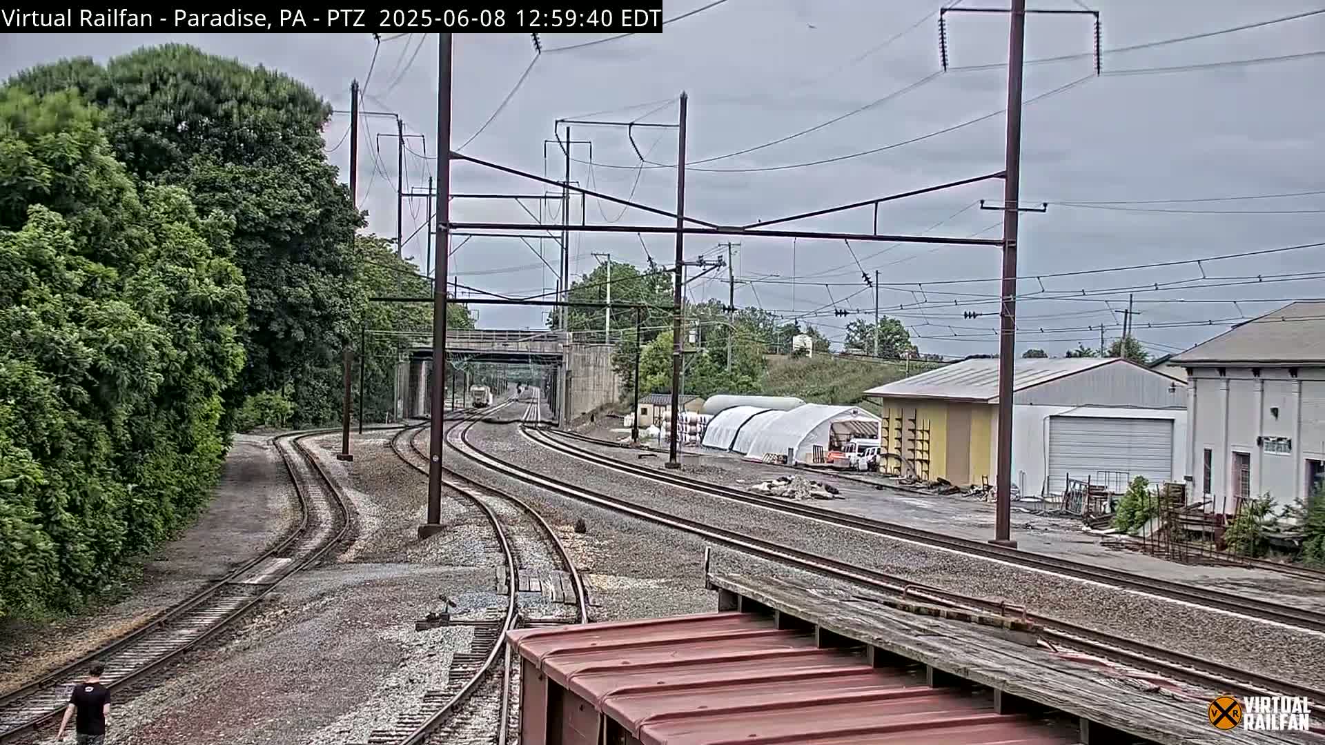 Several train tracks curve through a landscape of green trees and industrial buildings under an overcast sky.