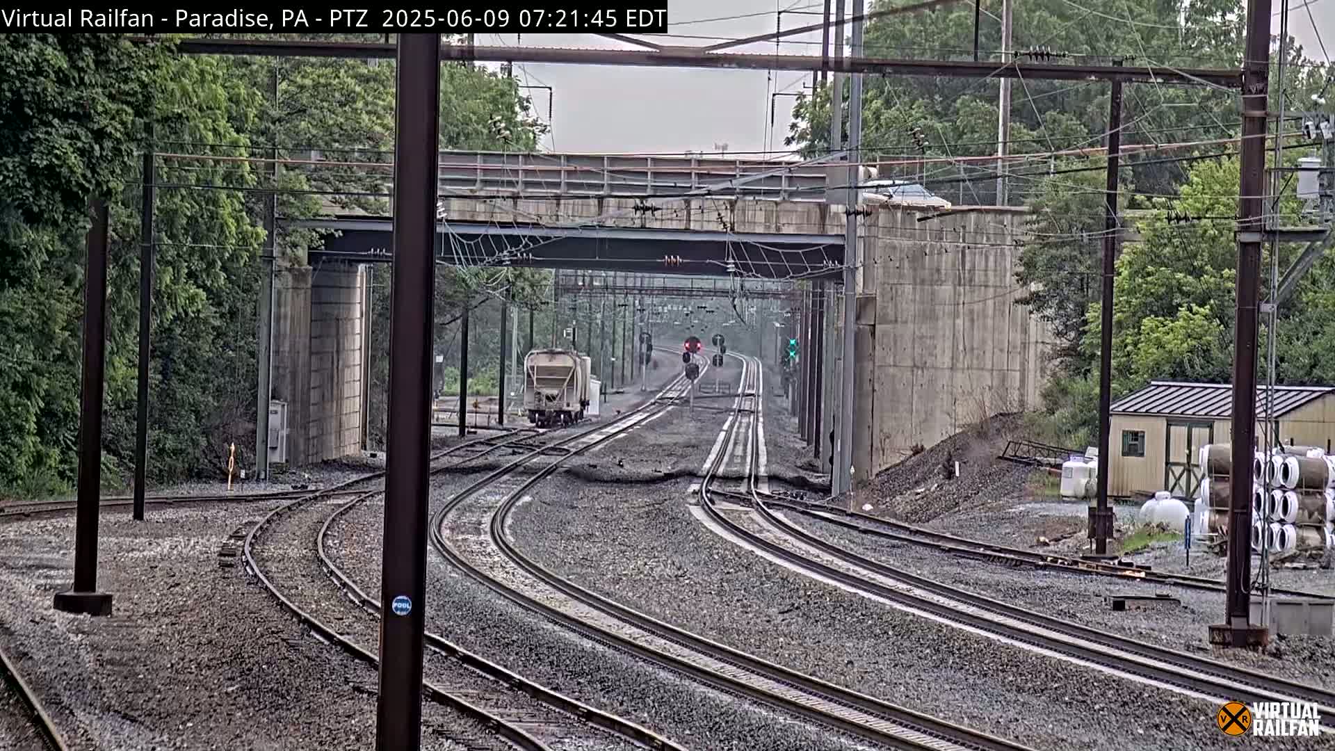 Multiple train tracks curve through a railway underpass under an overcast sky, with a light-colored rail car and a distant train visible.