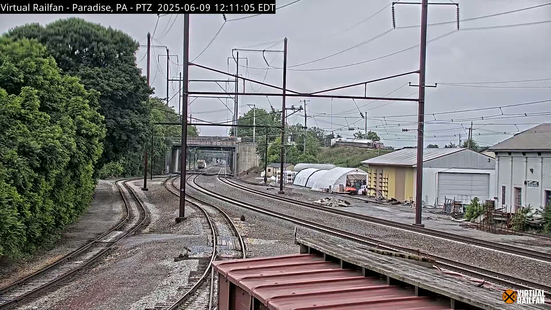Multiple sets of train tracks converge near a low-lying overpass under an overcast sky, with green trees and industrial buildings in the background.