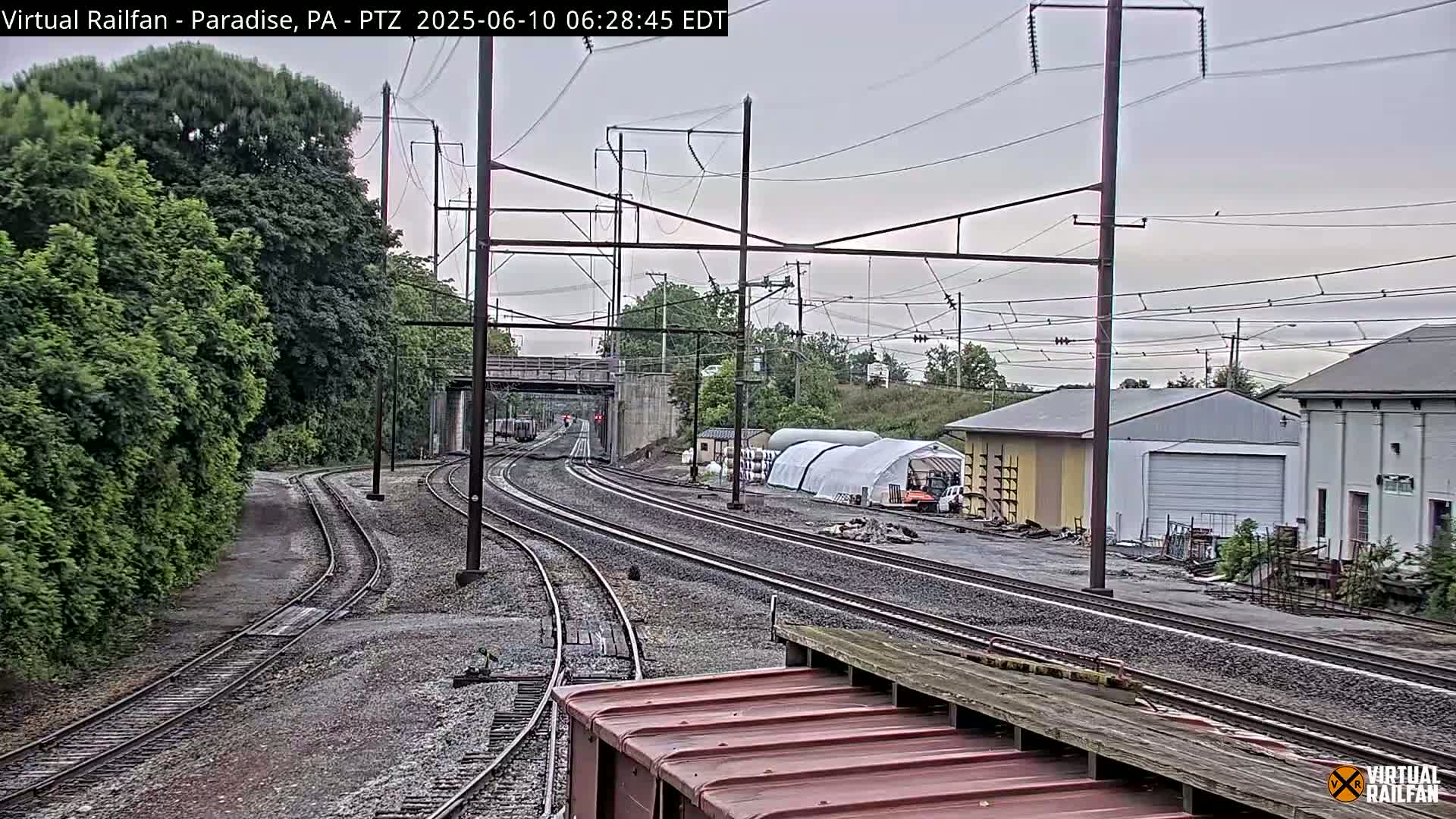 Several sets of train tracks diverge under an overcast sky, past lush green trees and industrial buildings.
