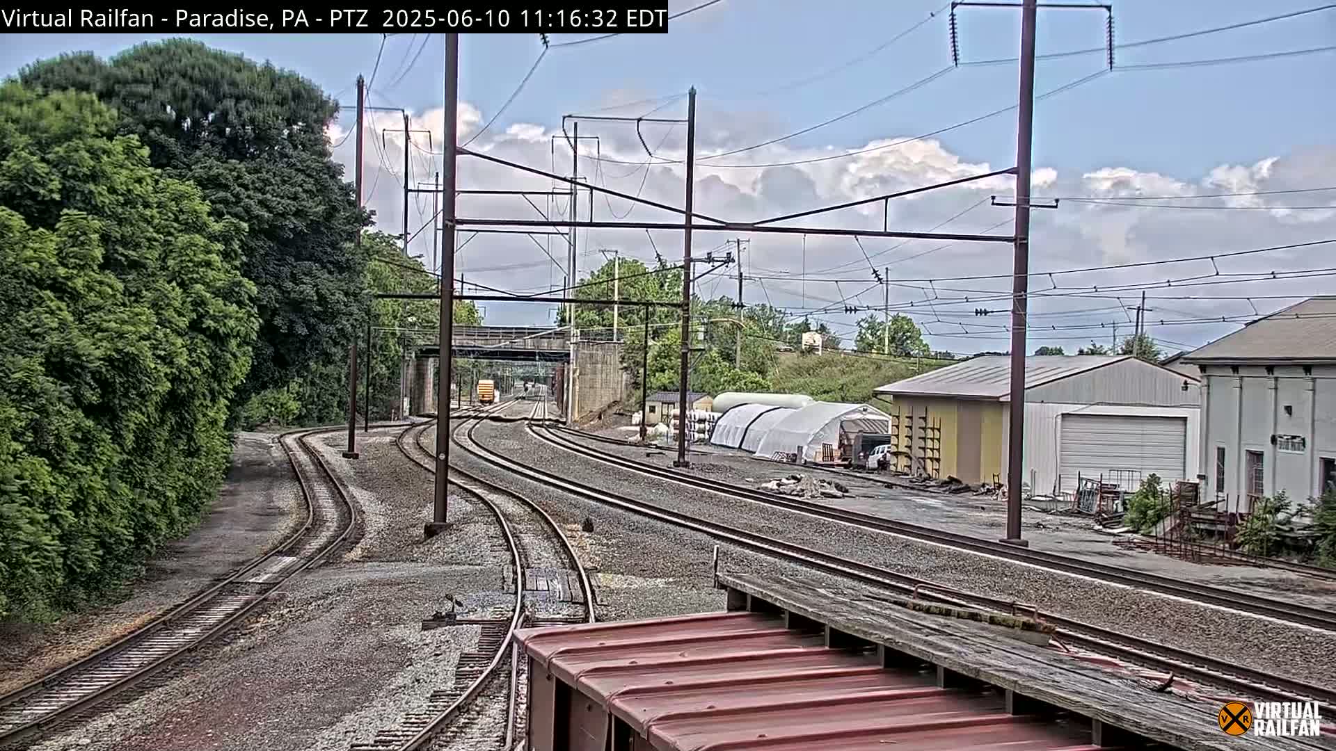 Converging railroad tracks under a partly cloudy sky pass by industrial buildings and lush green vegetation.
