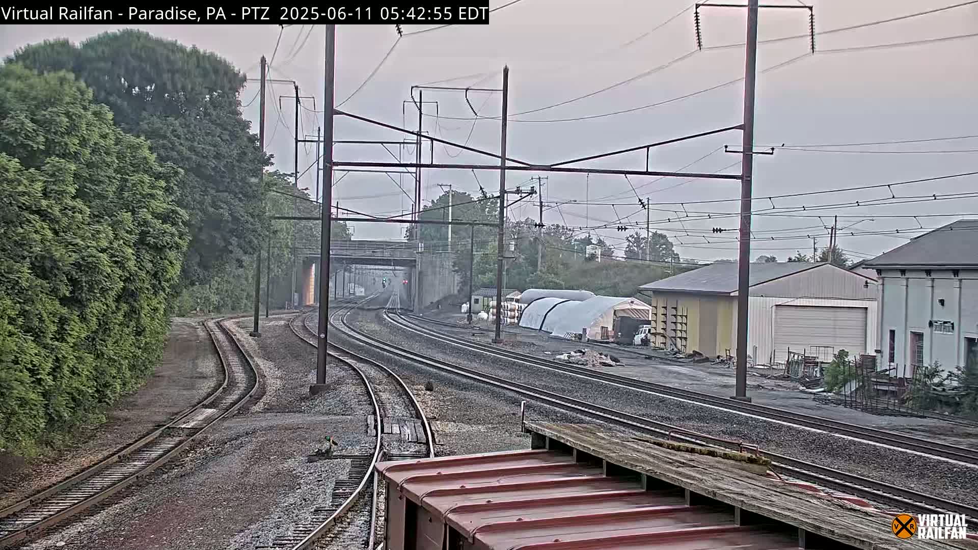 Several sets of train tracks curve through a landscape of trees, industrial buildings, and an overcast sky.