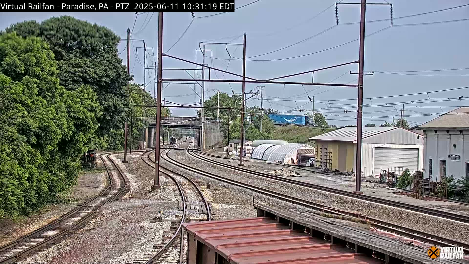Under a mostly clear sky, several sets of train tracks converge near a rail yard with some industrial buildings and green vegetation.