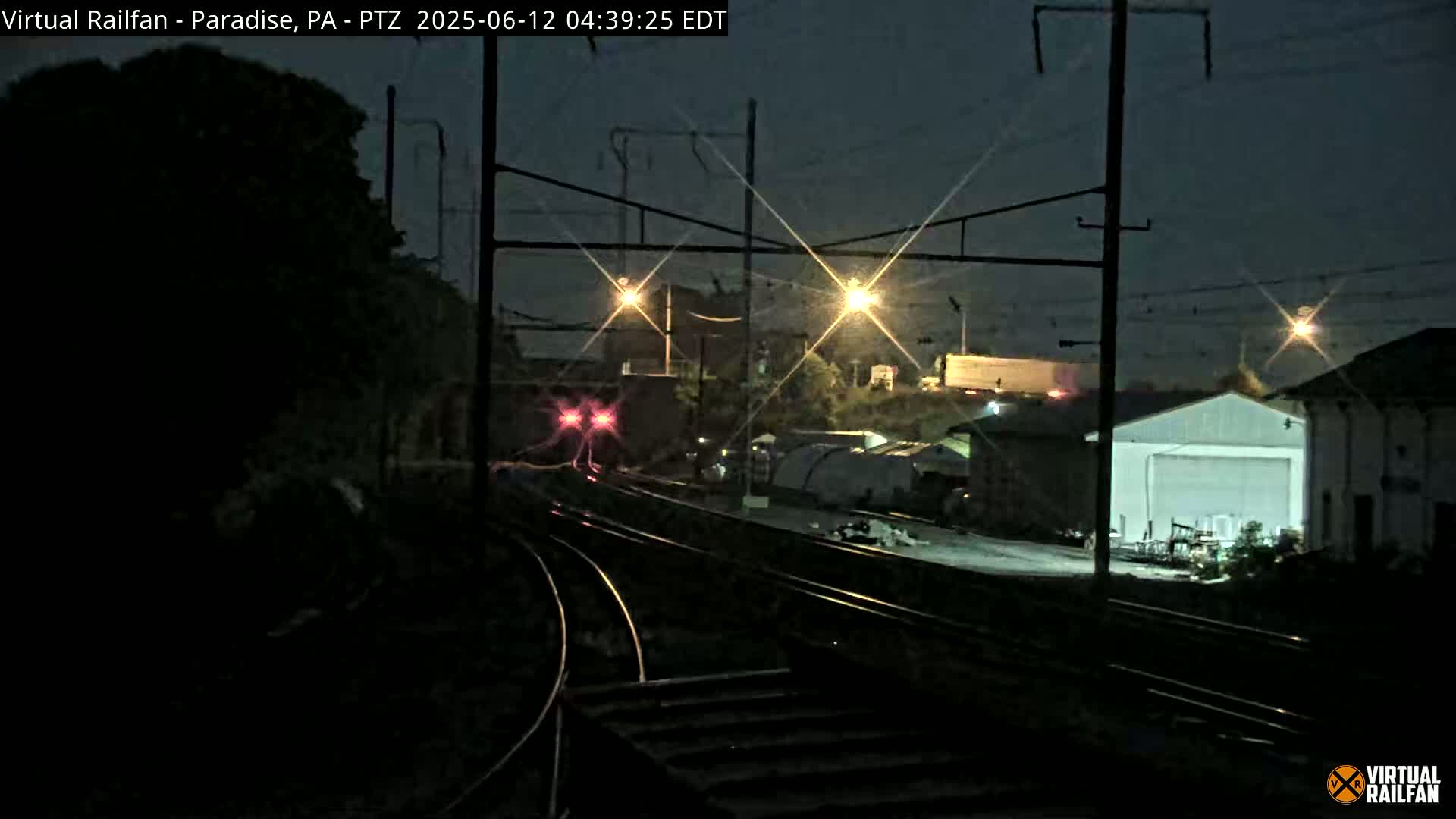 A nighttime view of railroad tracks with a train in the distance under a dark, possibly rainy sky, illuminated by streetlights and the train's lights.