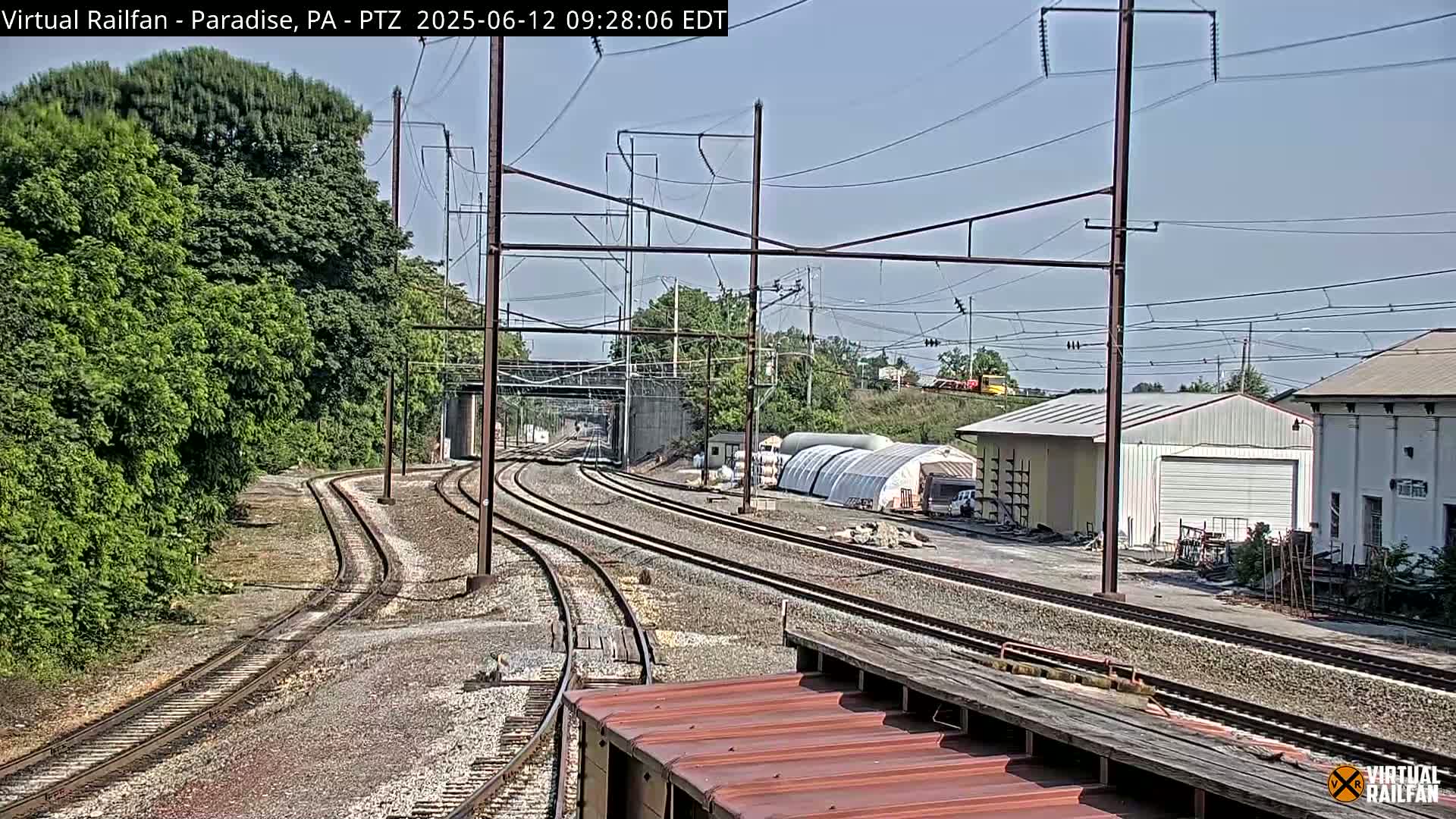 Converging railroad tracks under a mostly clear sky pass by a building and lush green foliage.