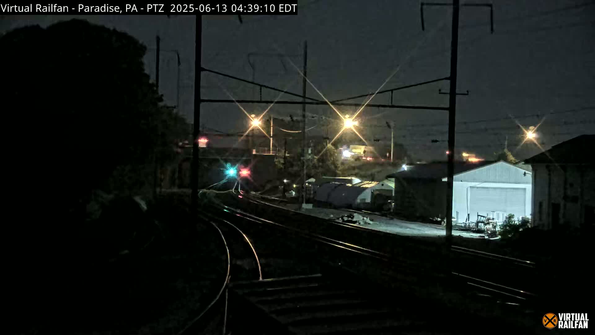 At night, under a dark sky, railroad tracks curve past a few buildings lit by streetlights.