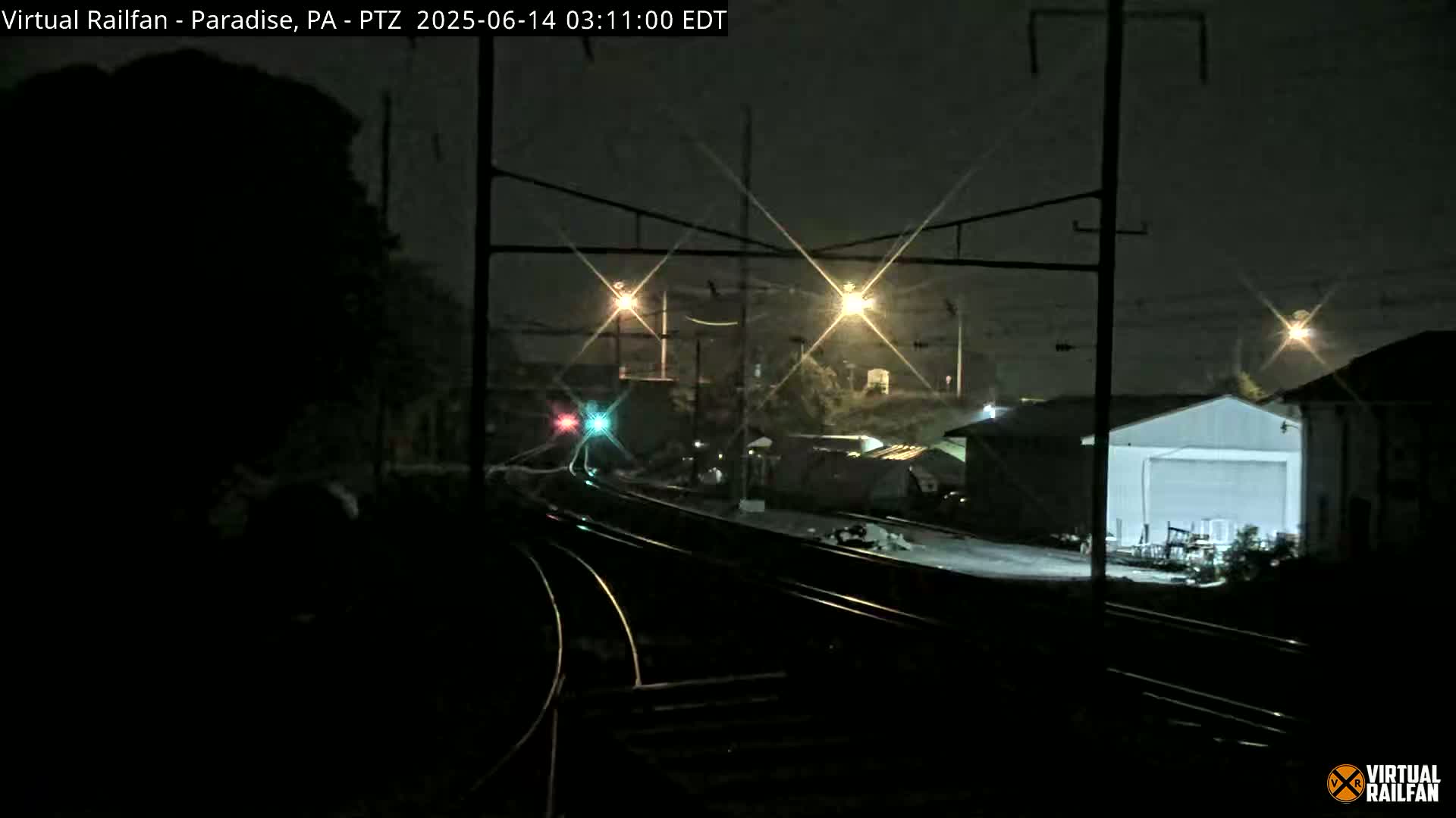 A nighttime view of railroad tracks and industrial buildings under a dark sky illuminated by several distant lights.