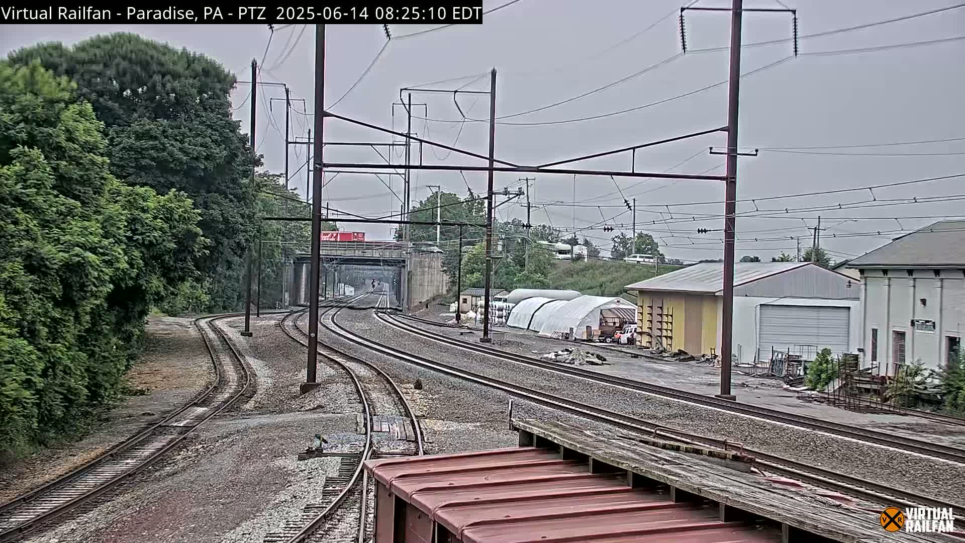 Multiple train tracks curve through a landscape of trees and industrial buildings under an overcast sky.