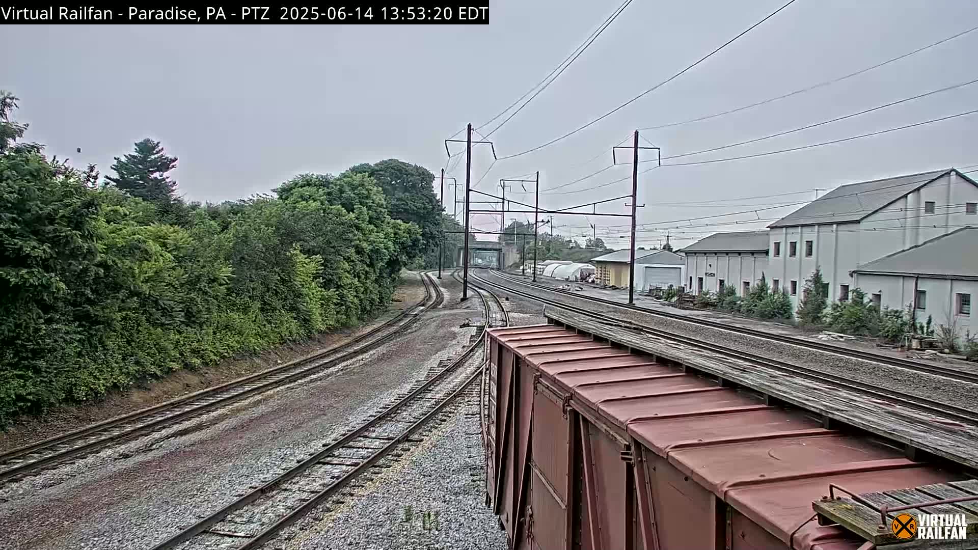 A view from atop a train car shows a railroad track splitting into two, leading to a row of industrial buildings under an overcast sky.