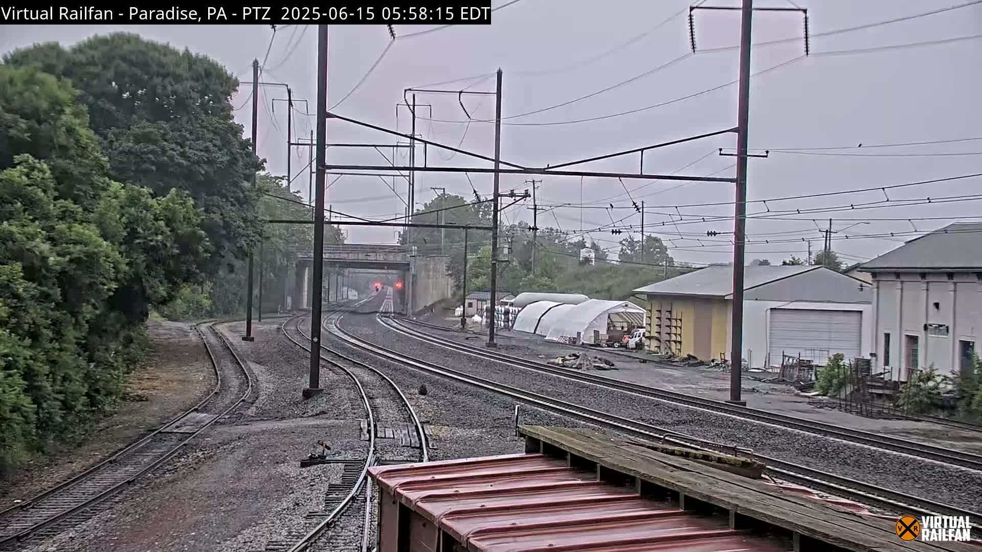 Multiple train tracks extend into the distance under an overcast sky, passing near a few buildings and lush green vegetation.