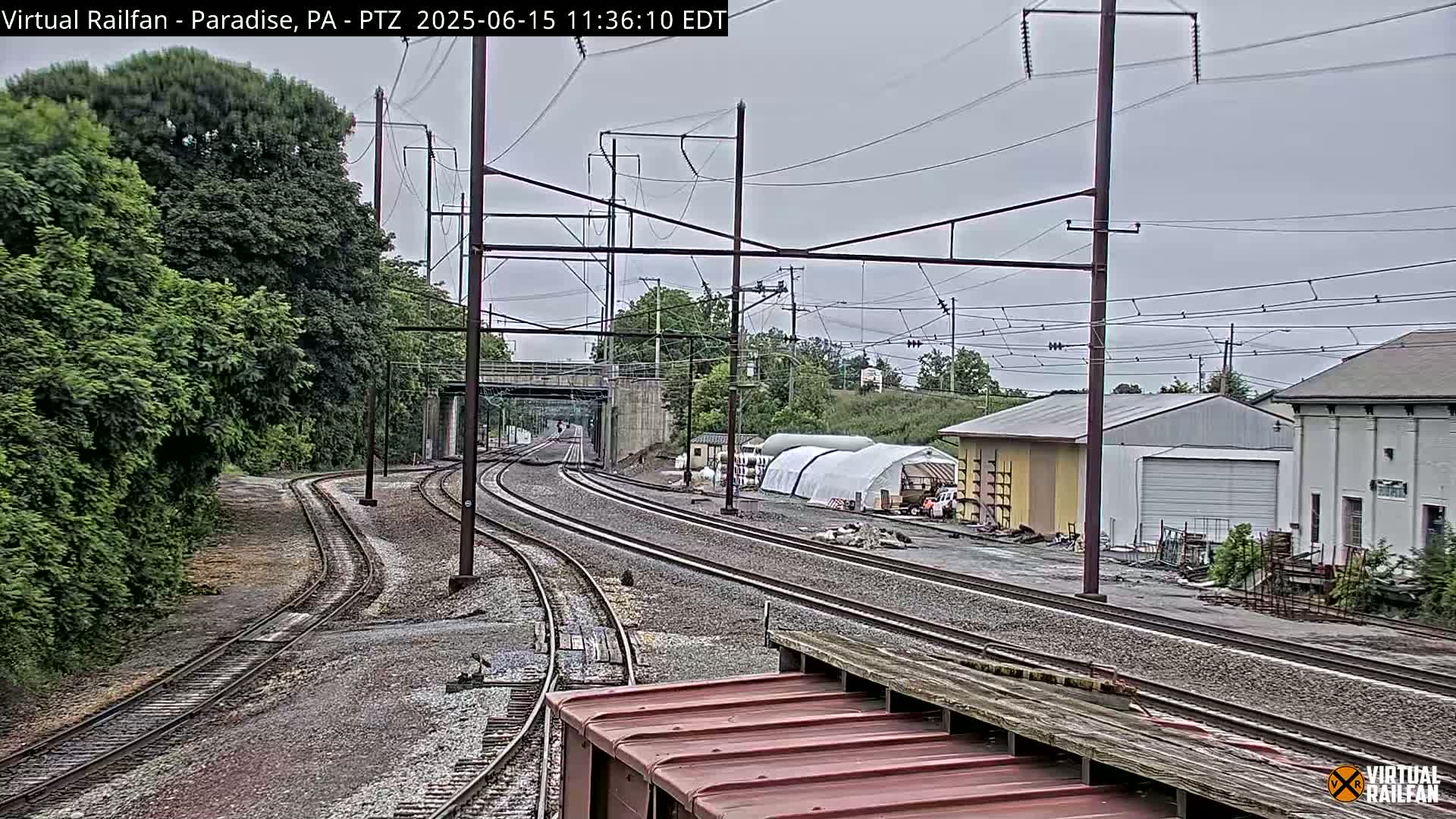 Multiple sets of train tracks curve through a landscape of green trees and industrial buildings under an overcast sky.