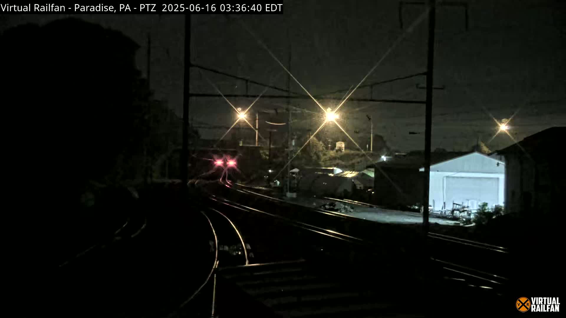 A nighttime view of railroad tracks with a train's lights visible in the distance, under a dark sky with some light rain or mist.