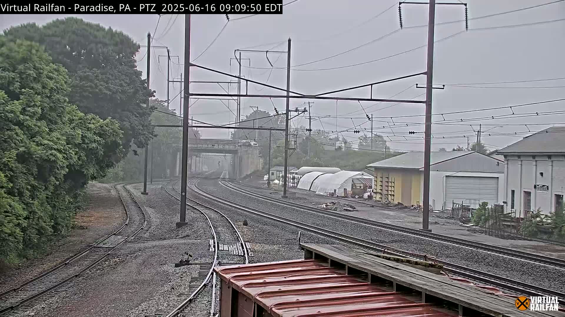 Multiple sets of train tracks extend into the distance under an overcast sky, past greenery, industrial buildings, and a portion of a red railroad car.