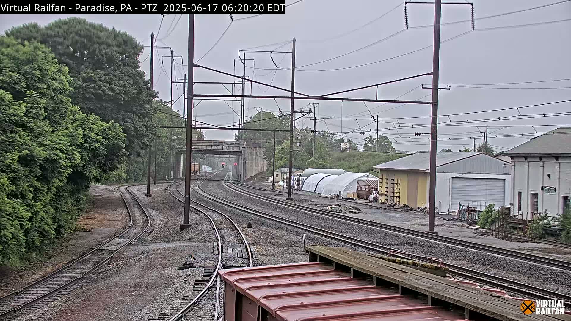 Multiple sets of train tracks curve through a landscape of green trees and industrial buildings under an overcast sky.