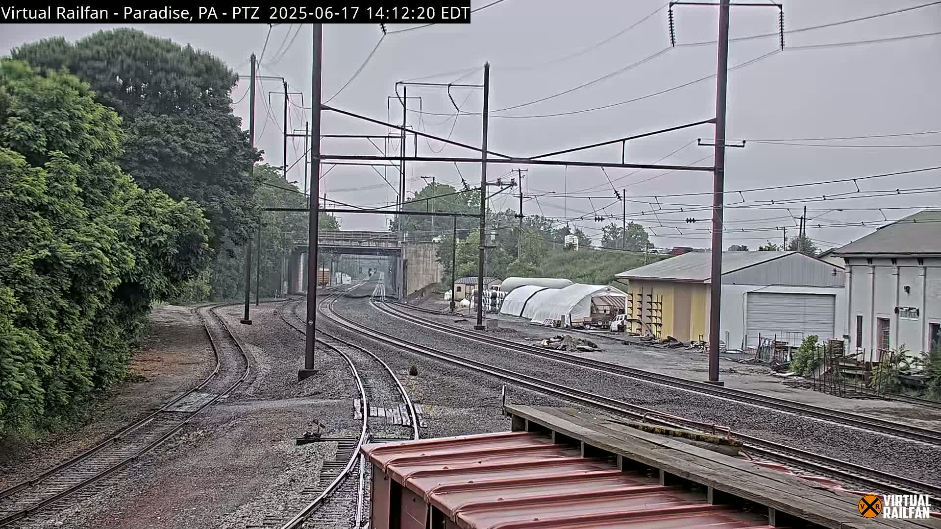 Converging railroad tracks extend toward a bridge and industrial buildings under an overcast sky.