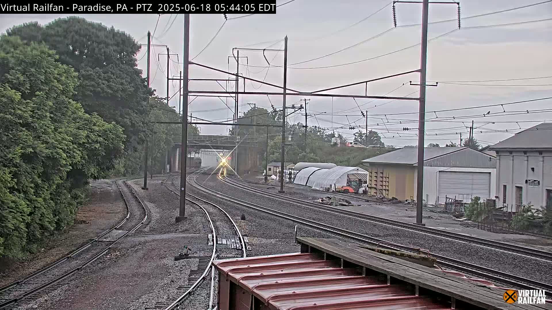 A train passes under an overpass on a cloudy day, with railroad tracks stretching into the distance past industrial buildings and lush green trees.