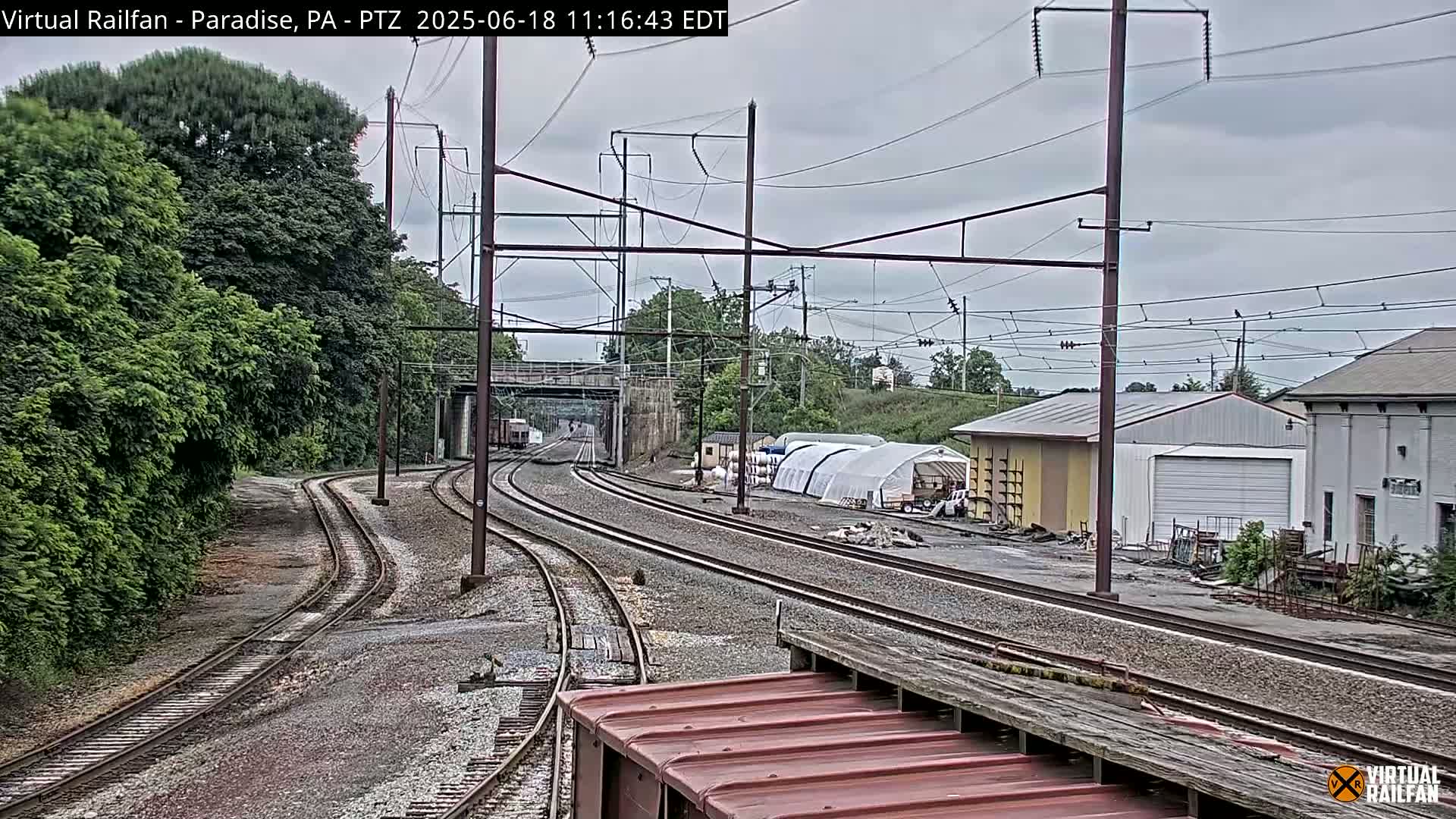 Multiple sets of train tracks curve through a landscape of trees, industrial buildings, and overcast skies.