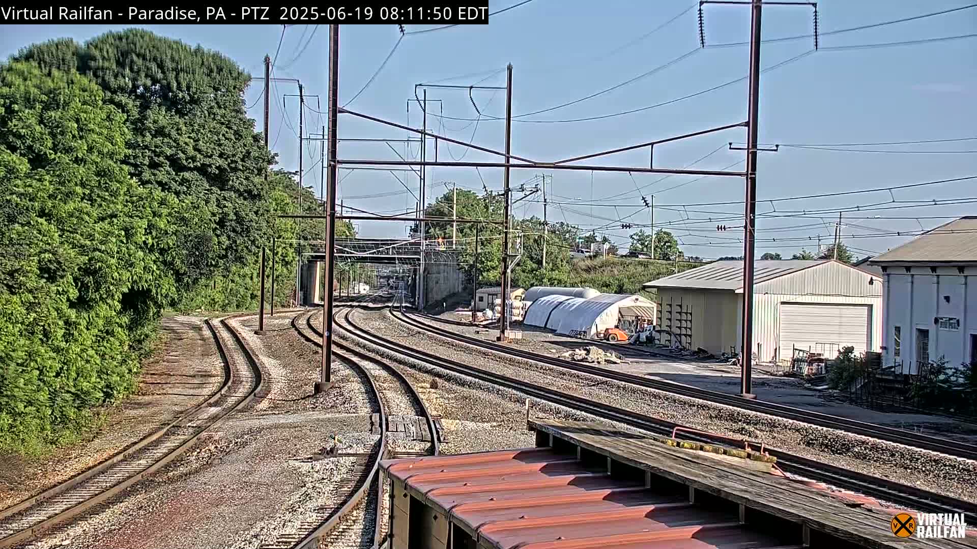 Converging train tracks on a sunny day pass by a lush green hillside, industrial buildings, and a large white temporary structure.