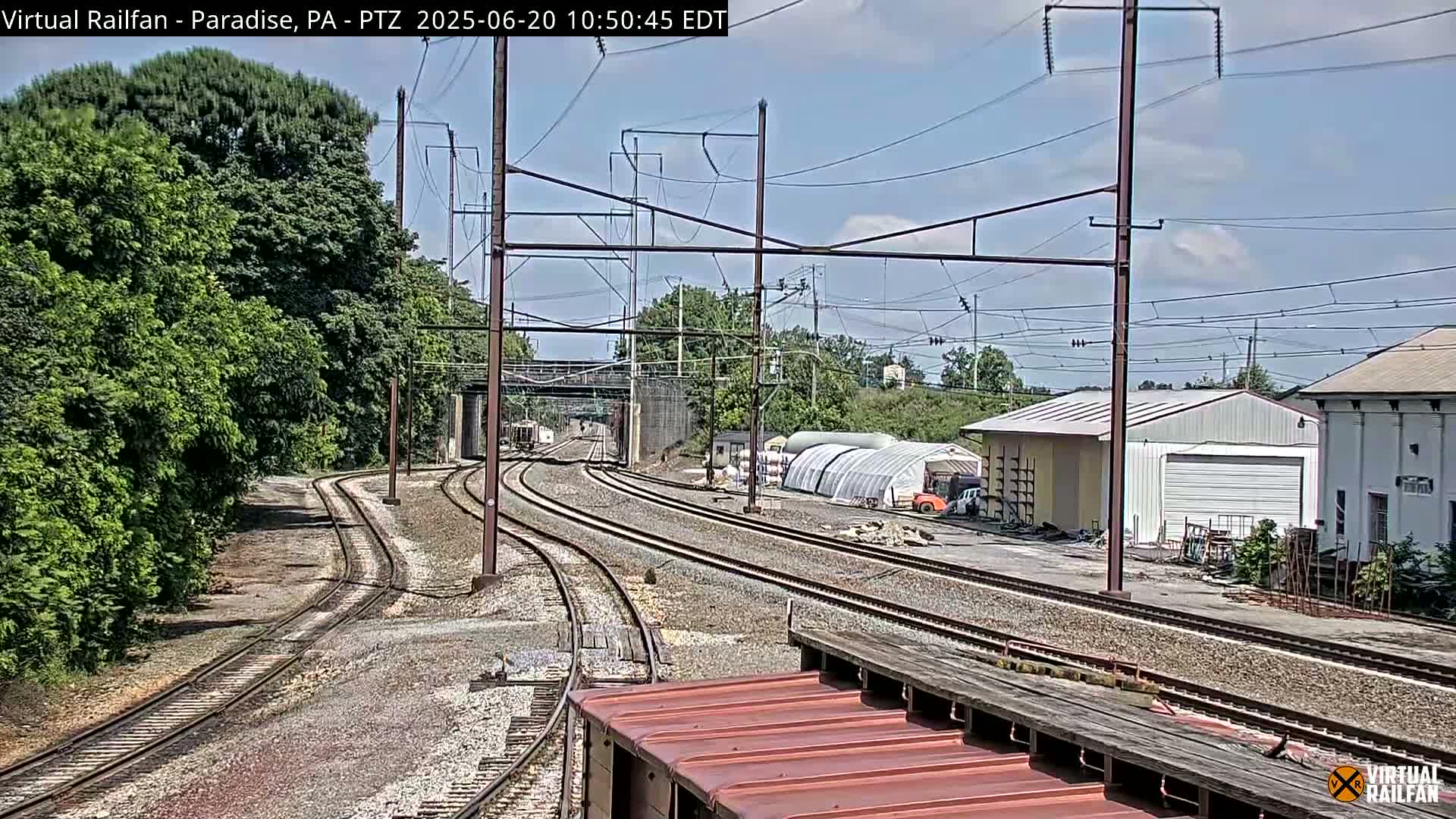 Multiple train tracks curve through a landscape of green trees, industrial buildings, and a partially visible greenhouse under a partly cloudy sky.
