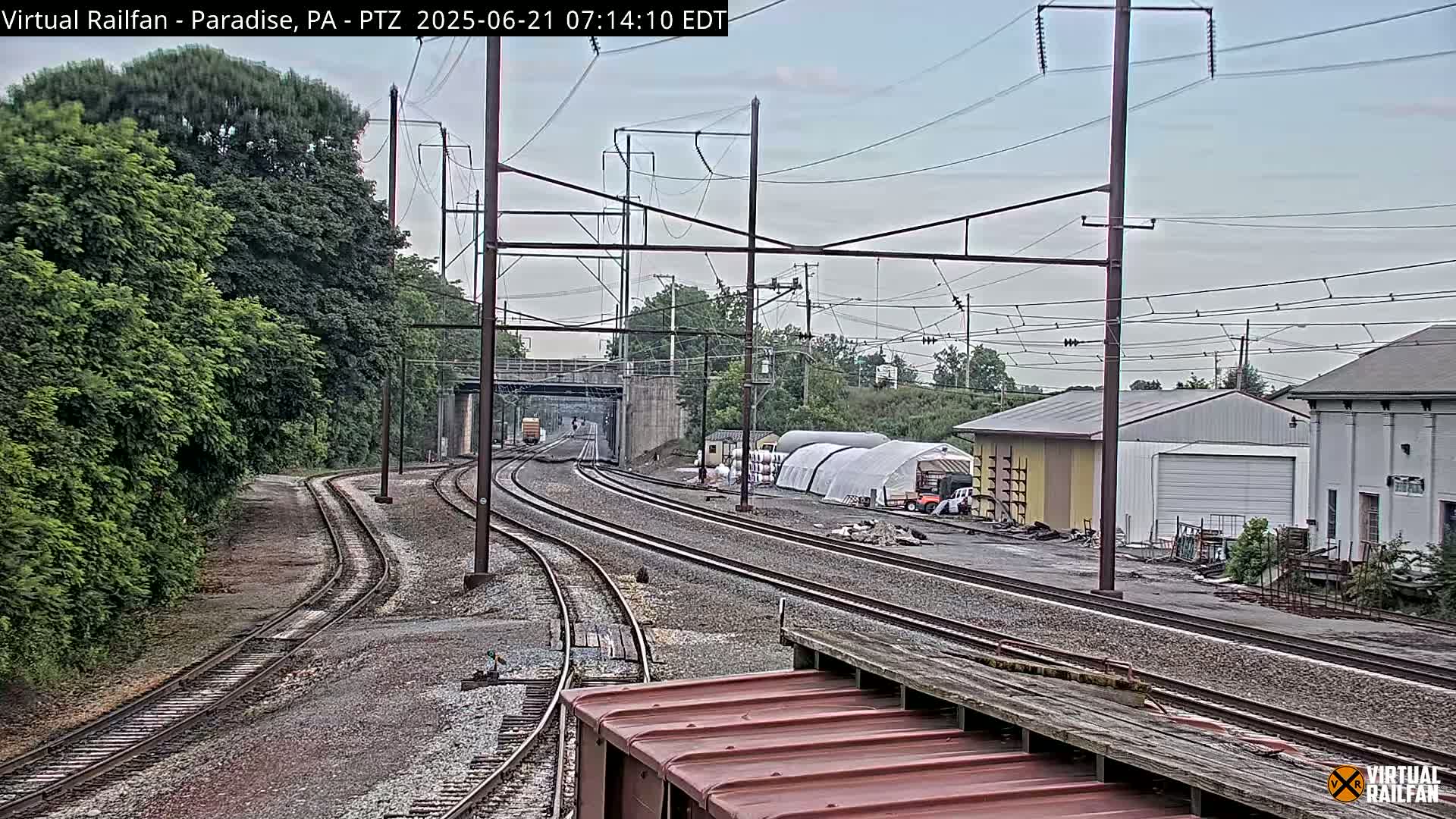 Several train tracks curve through a landscape of green trees and industrial buildings under an overcast sky.