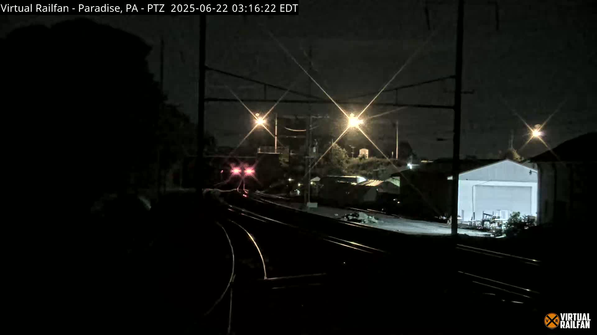 A nighttime view of a train passing by railroad tracks, with several buildings and streetlights visible in the background under a dark sky.