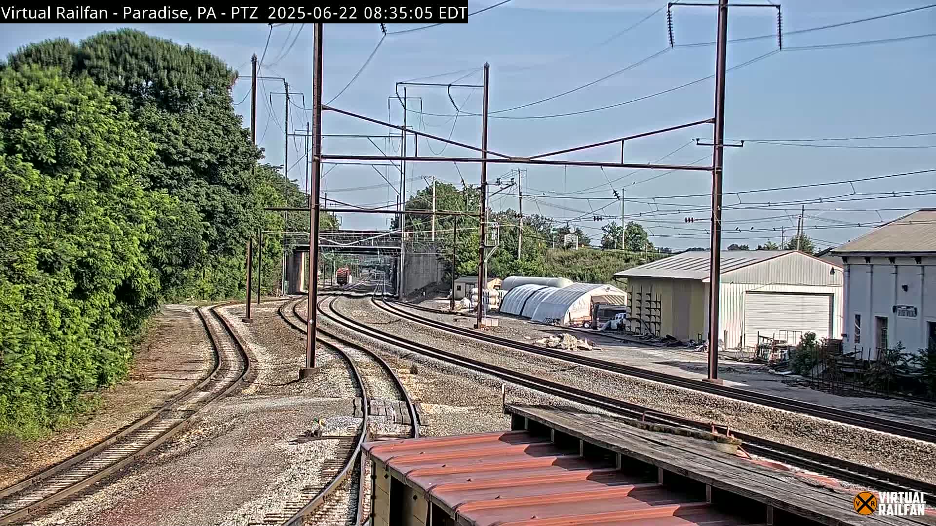 Several sets of train tracks curve away from the camera, passing alongside a lush green embankment, industrial buildings, and a large, partially visible shed under a mostly clear sky.