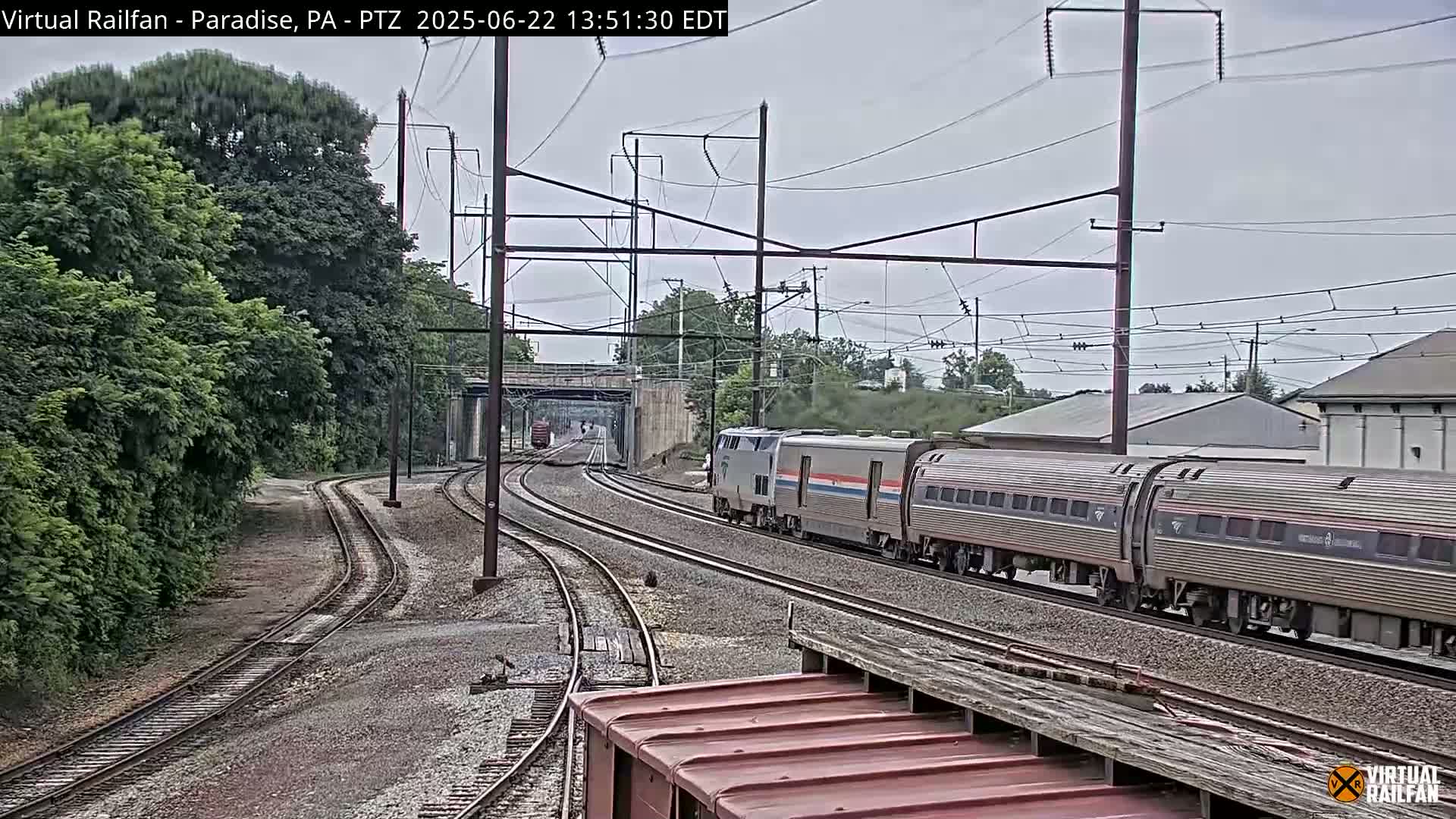 A long passenger train travels along a railway track under an overcast sky, passing alongside lush green trees and a low building.