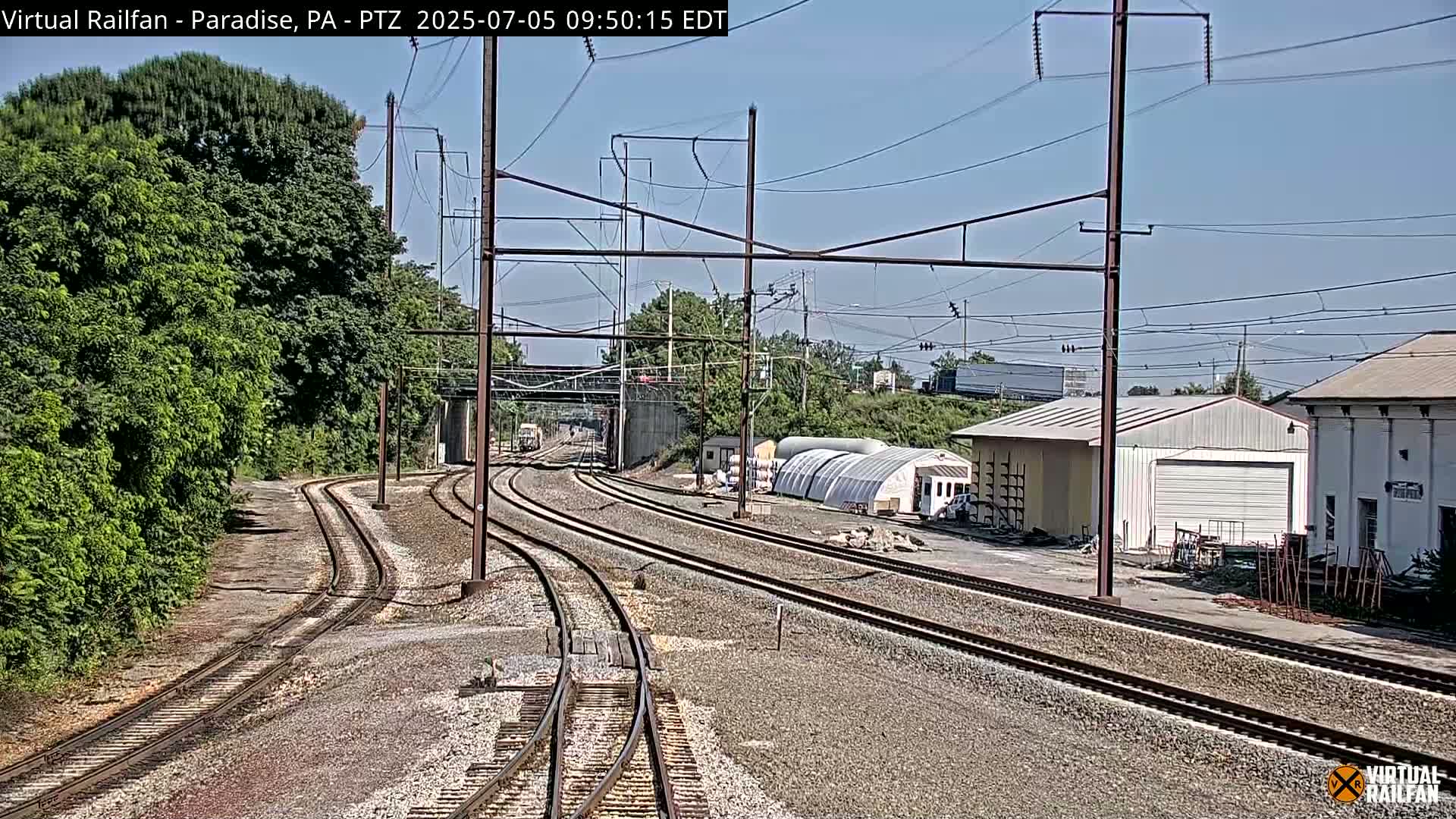 Multiple sets of train tracks curve toward a distant bridge under a clear, sunny sky, alongside lush green trees and industrial buildings.