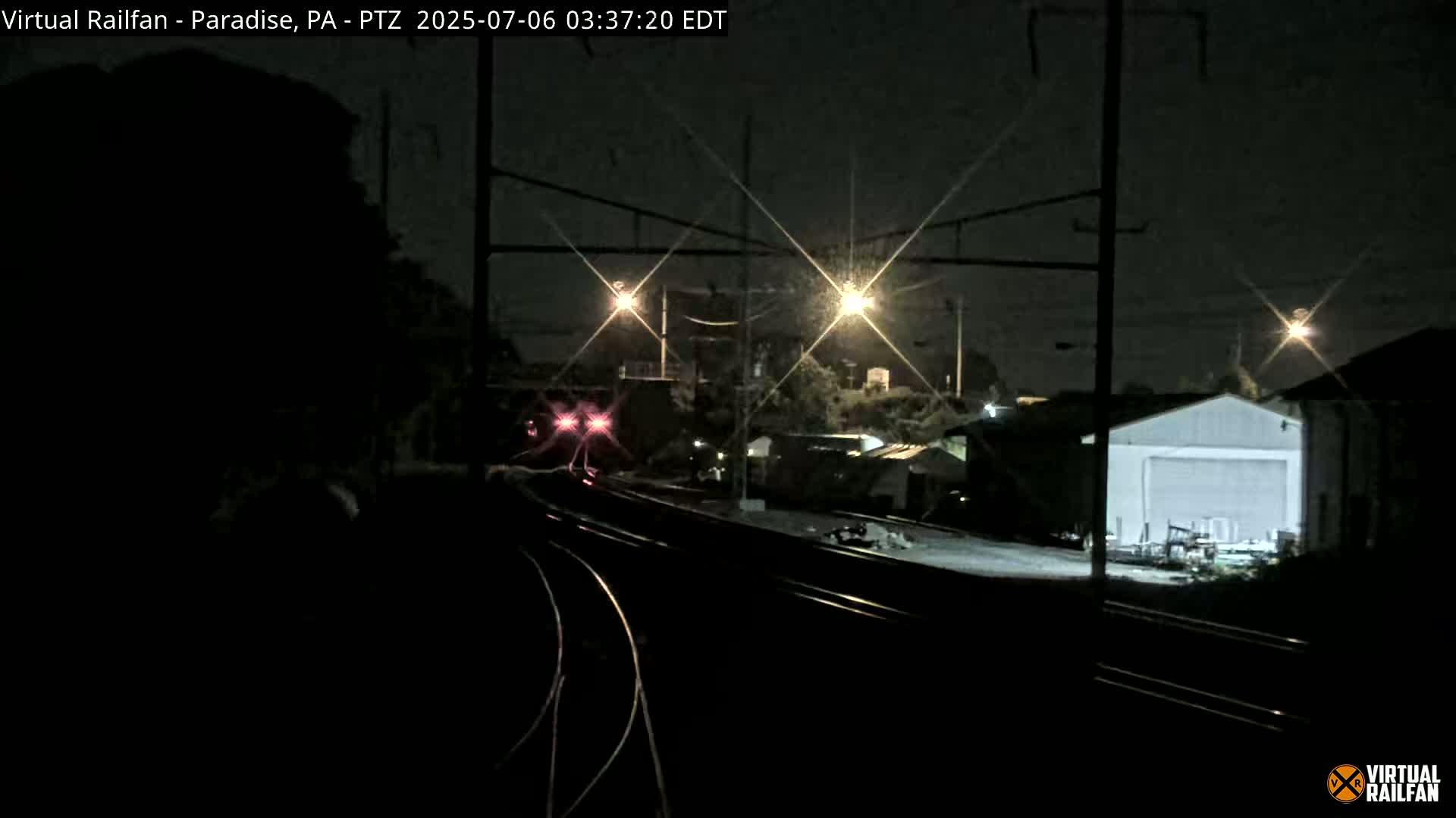 A train's rear red lights are visible at night as it approaches a rail yard illuminated by several streetlights.