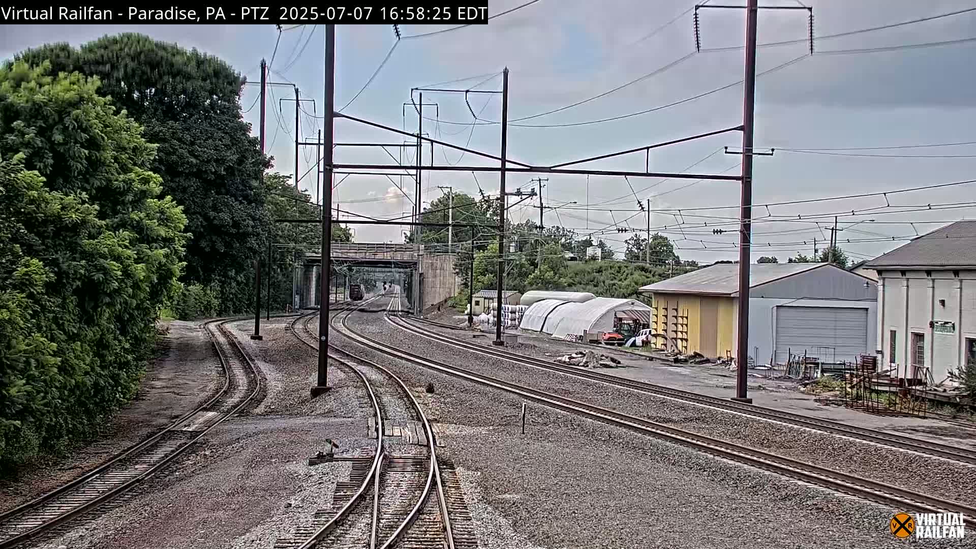 Multiple train tracks curve toward a distant bridge under an overcast sky, passing alongside buildings and lush green vegetation.