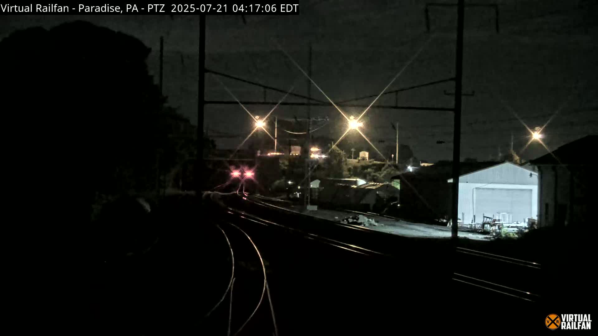 A nighttime view of railroad tracks, with a train in the distance and industrial buildings visible under bright lights.