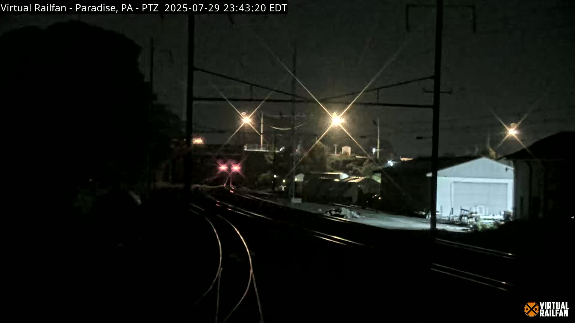 A nighttime view of railroad tracks, with a train in the distance and industrial buildings visible under bright lights.