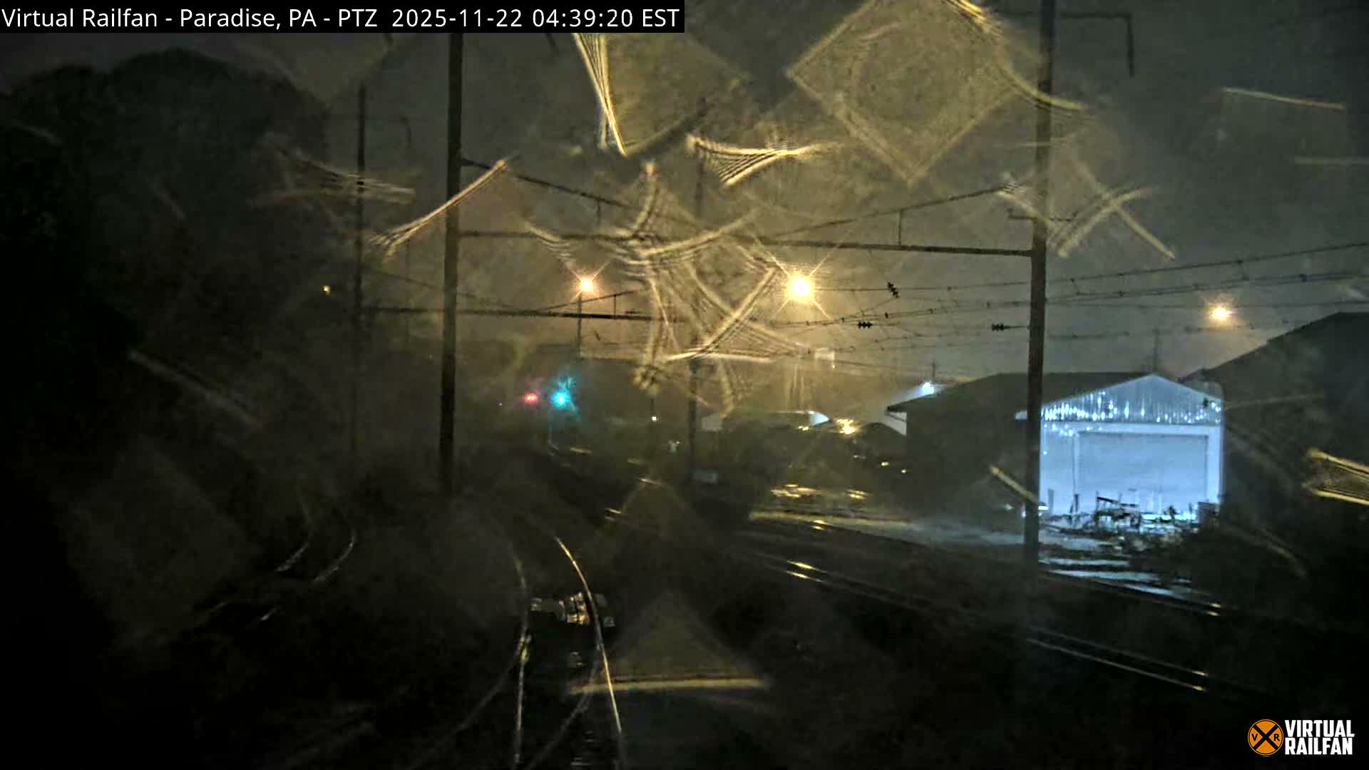 A dark, rainy night captures multiple train tracks and overhead catenary lines, with streetlights creating prominent starburst flares across the camera lens, alongside a green and red traffic light and buildings in the distance.