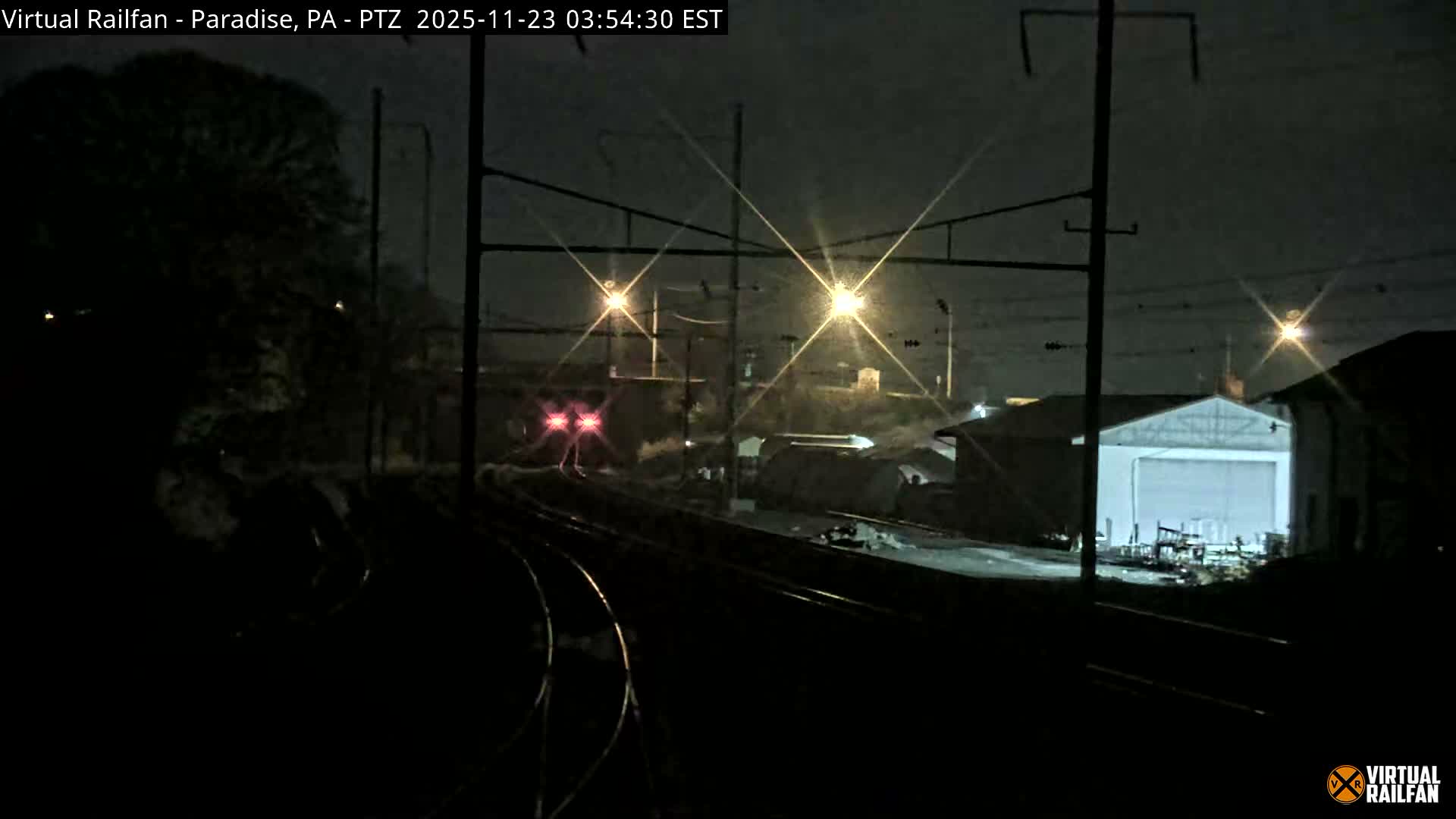 A nighttime view reveals multiple curving railway tracks, industrial buildings, and bright streetlights with prominent lens flares under a clear dark sky.