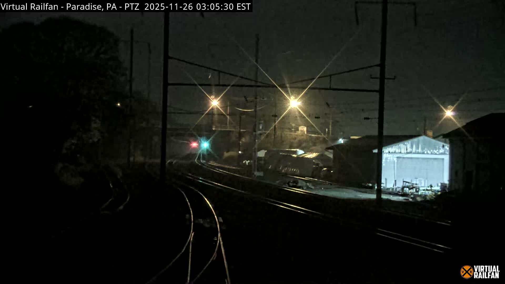 At night, multiple train tracks curve past industrial buildings, illuminated by overhead streetlights and prominent red and green signal lights, under a clear sky.