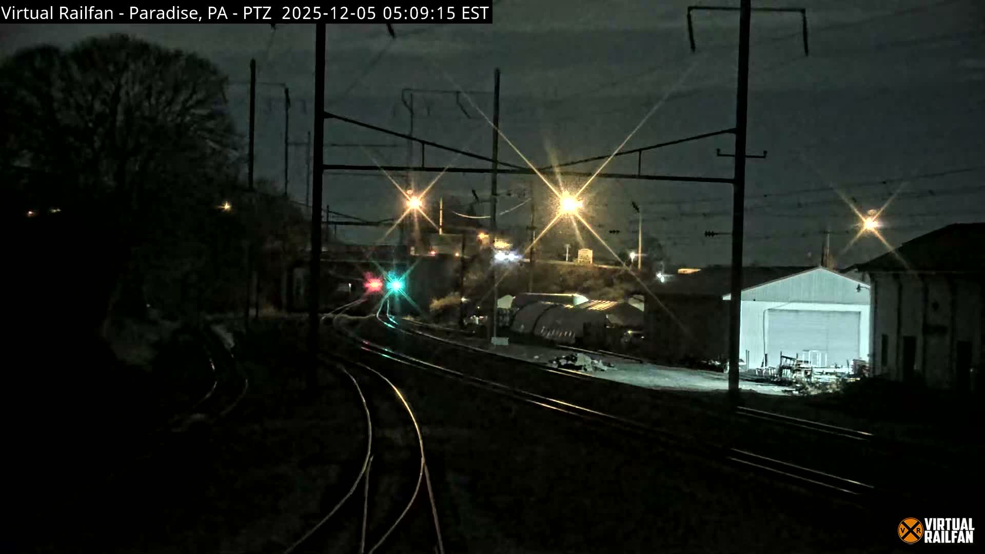 Multiple train tracks curve through an industrial area under a clear, dark night sky, illuminated by bright overhead lights and a red and green railway signal.