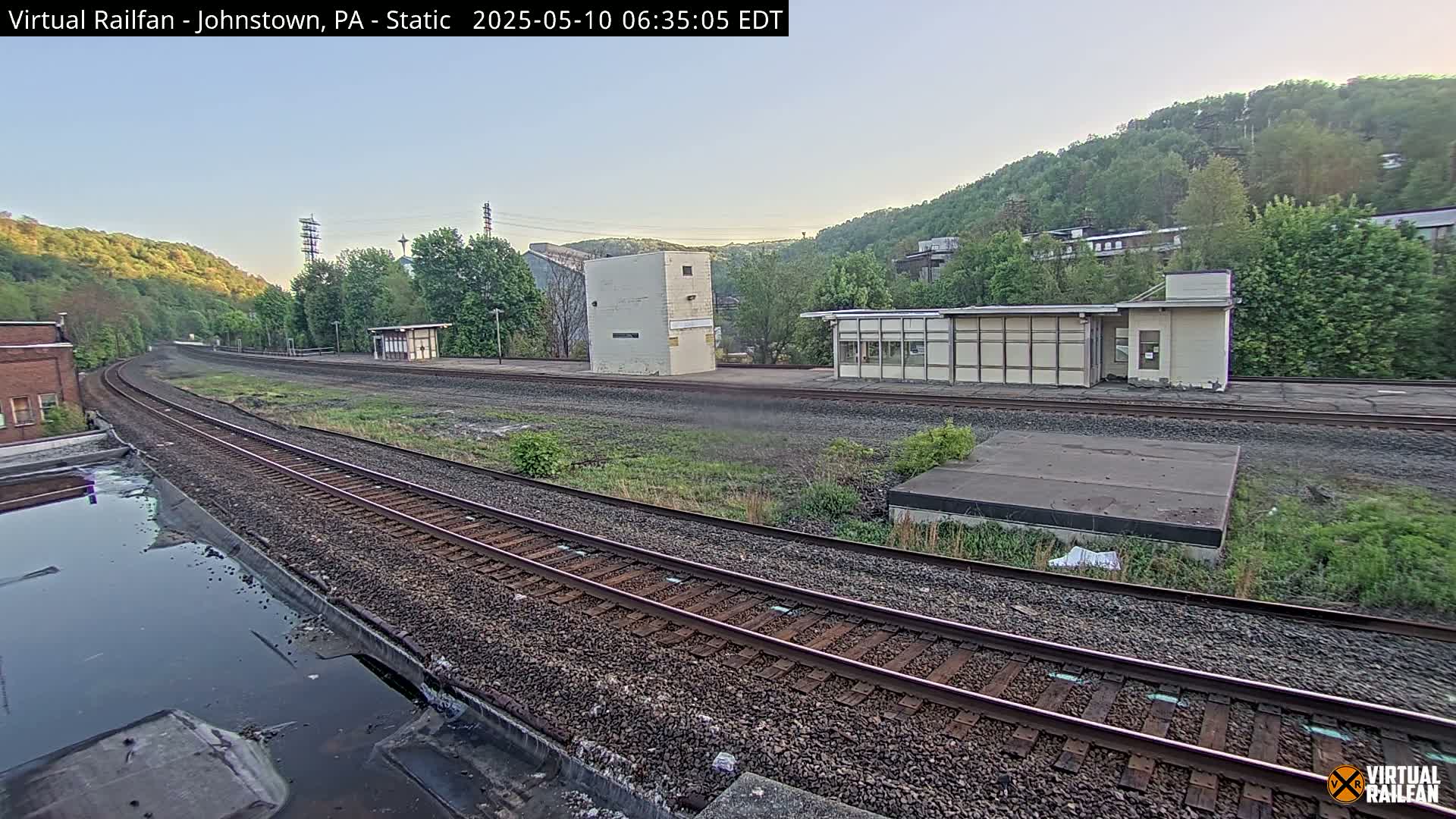 Train tracks curve through a mostly-clear, early morning landscape, past several small buildings and a hillside covered in green trees.