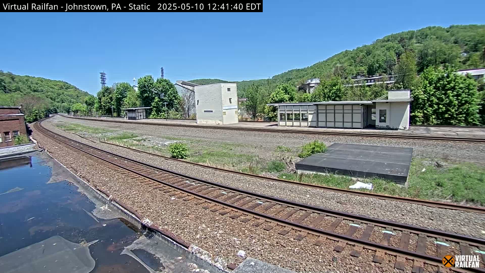 A curving railway track winds through a sunny landscape, past a small building and a larger structure, with lush green hills in the background.