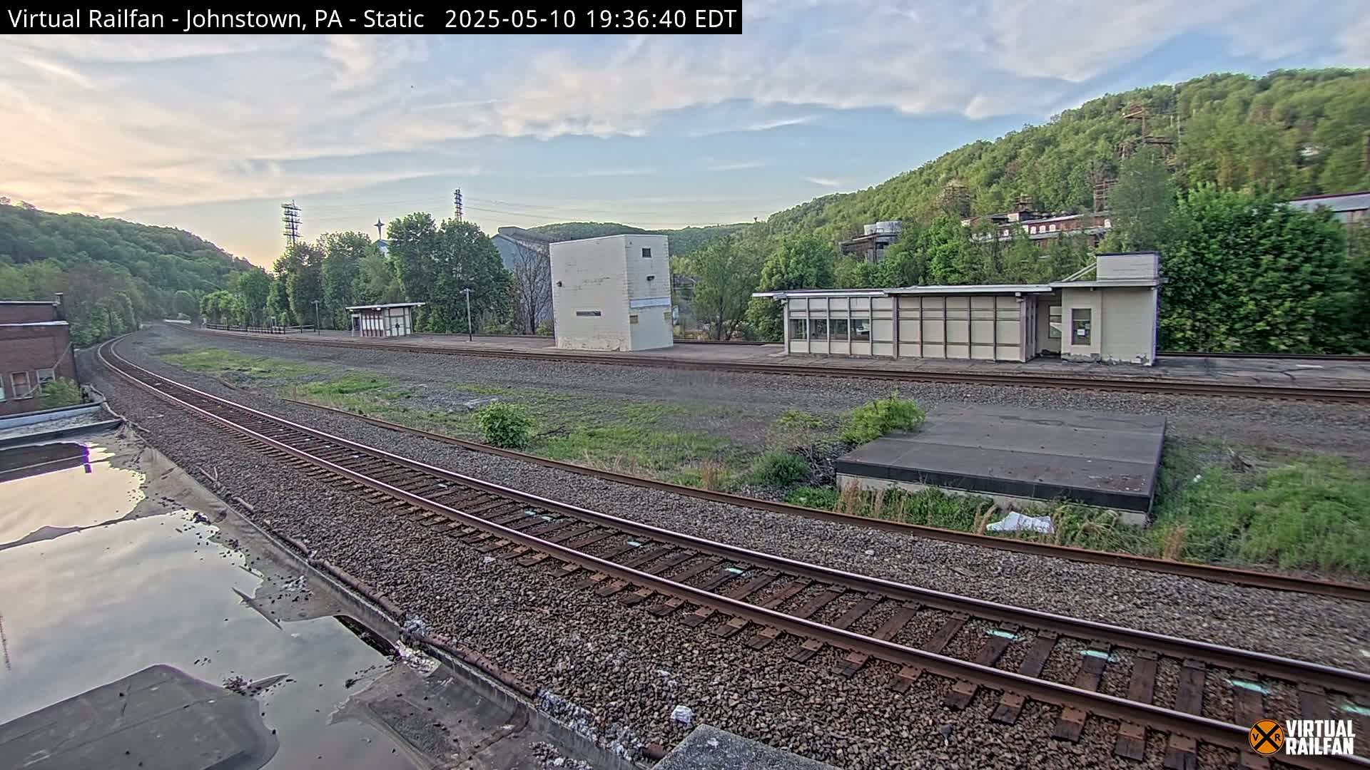 A railway track curves through a mostly clear, late-afternoon scene, passing by several small buildings and a hillside covered in green trees.