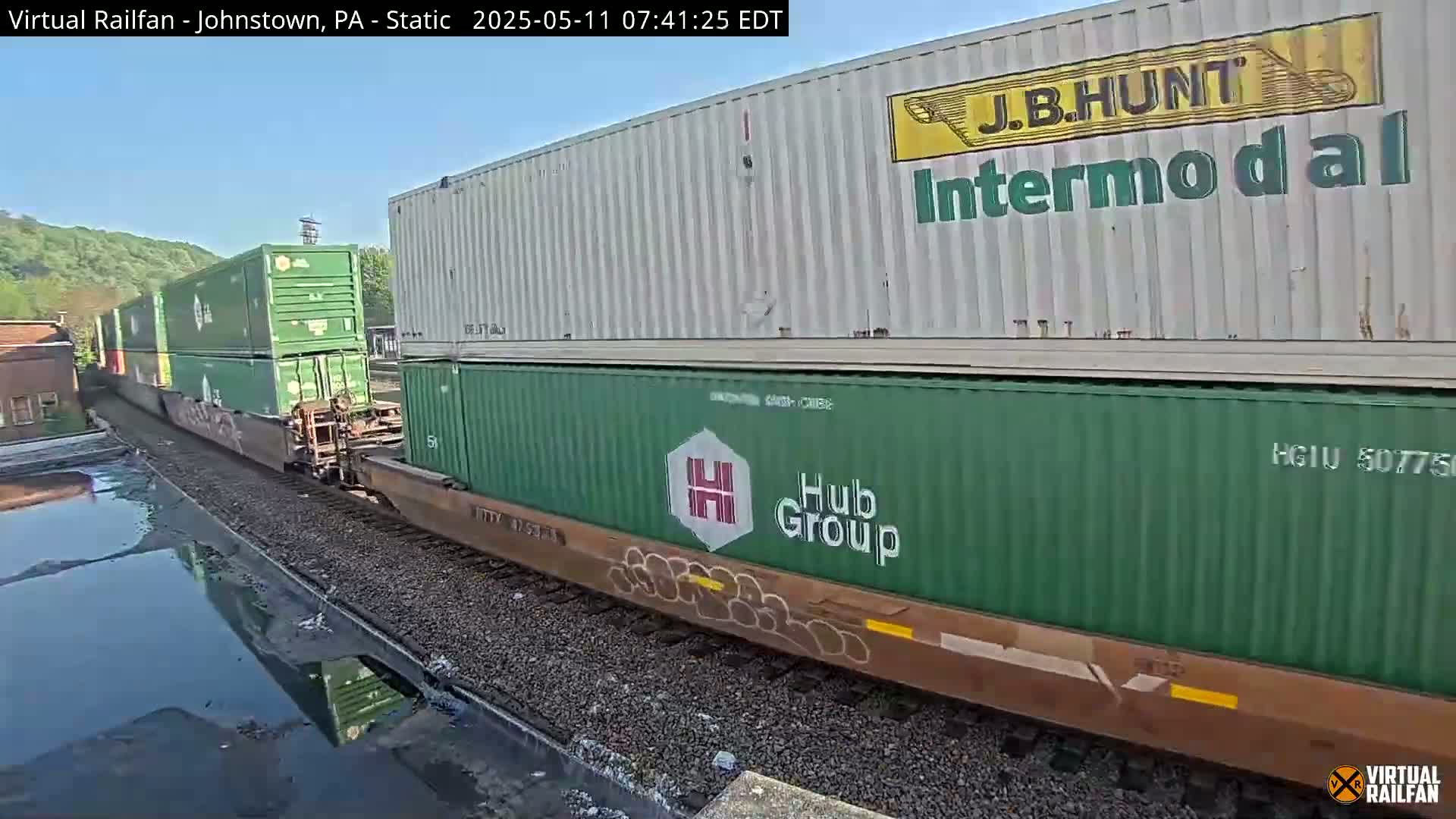 A freight train carrying various shipping containers moves along railroad tracks on a partly sunny day.