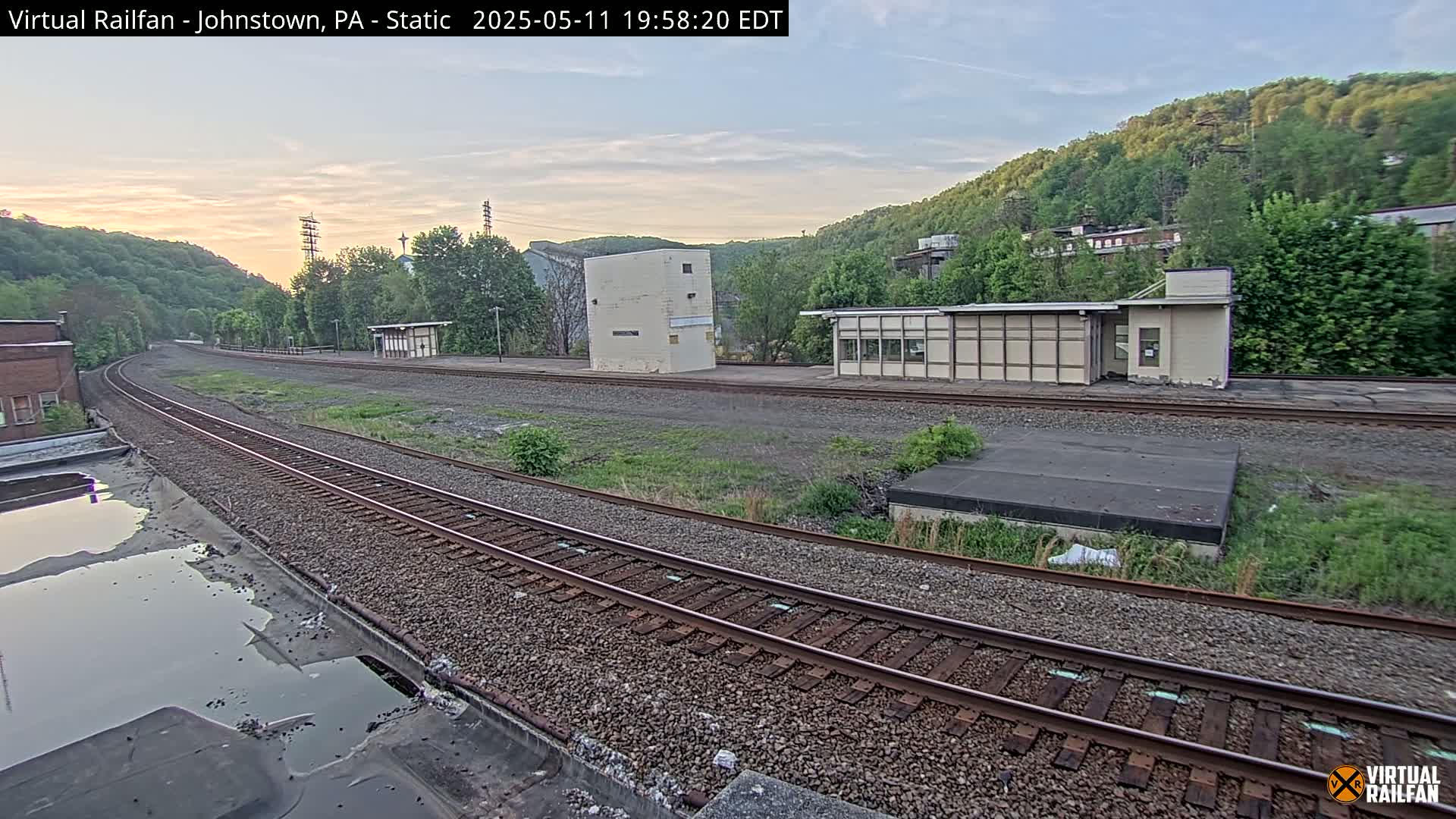 A train track curves gently through a rail yard with several small buildings under a twilight sky.