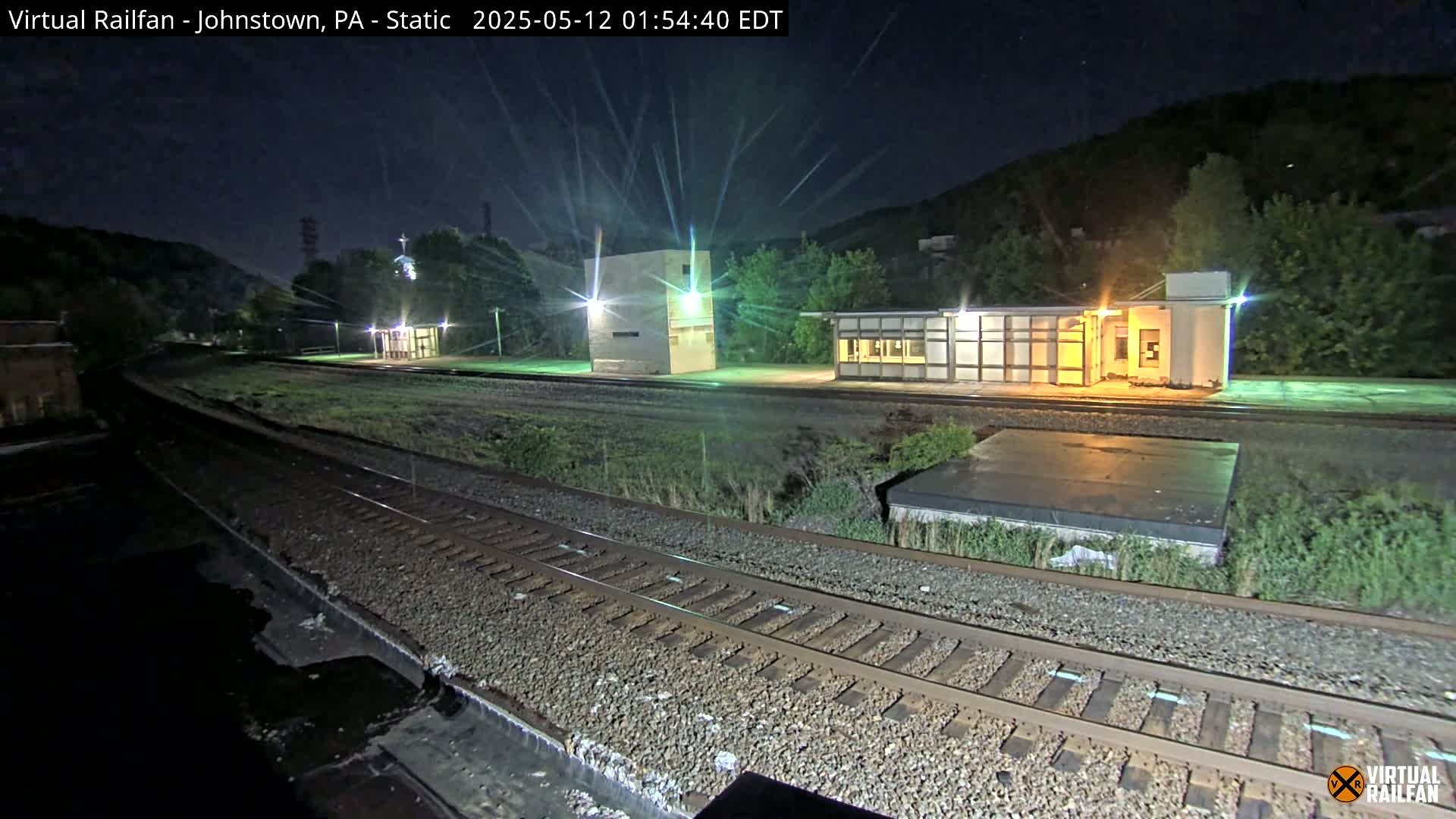A nighttime view of a train station with several buildings and railroad tracks under a clear, starry sky.