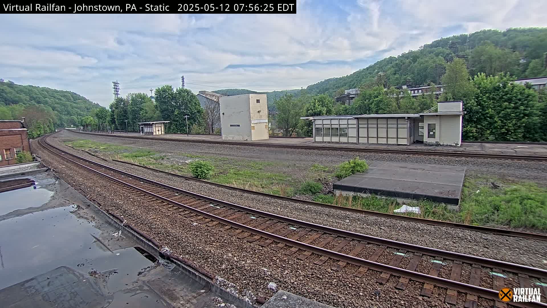 A mostly cloudy day reveals a railway scene with train tracks curving through a landscape of overgrown vegetation, several low buildings, and hills covered in lush green trees in the background.