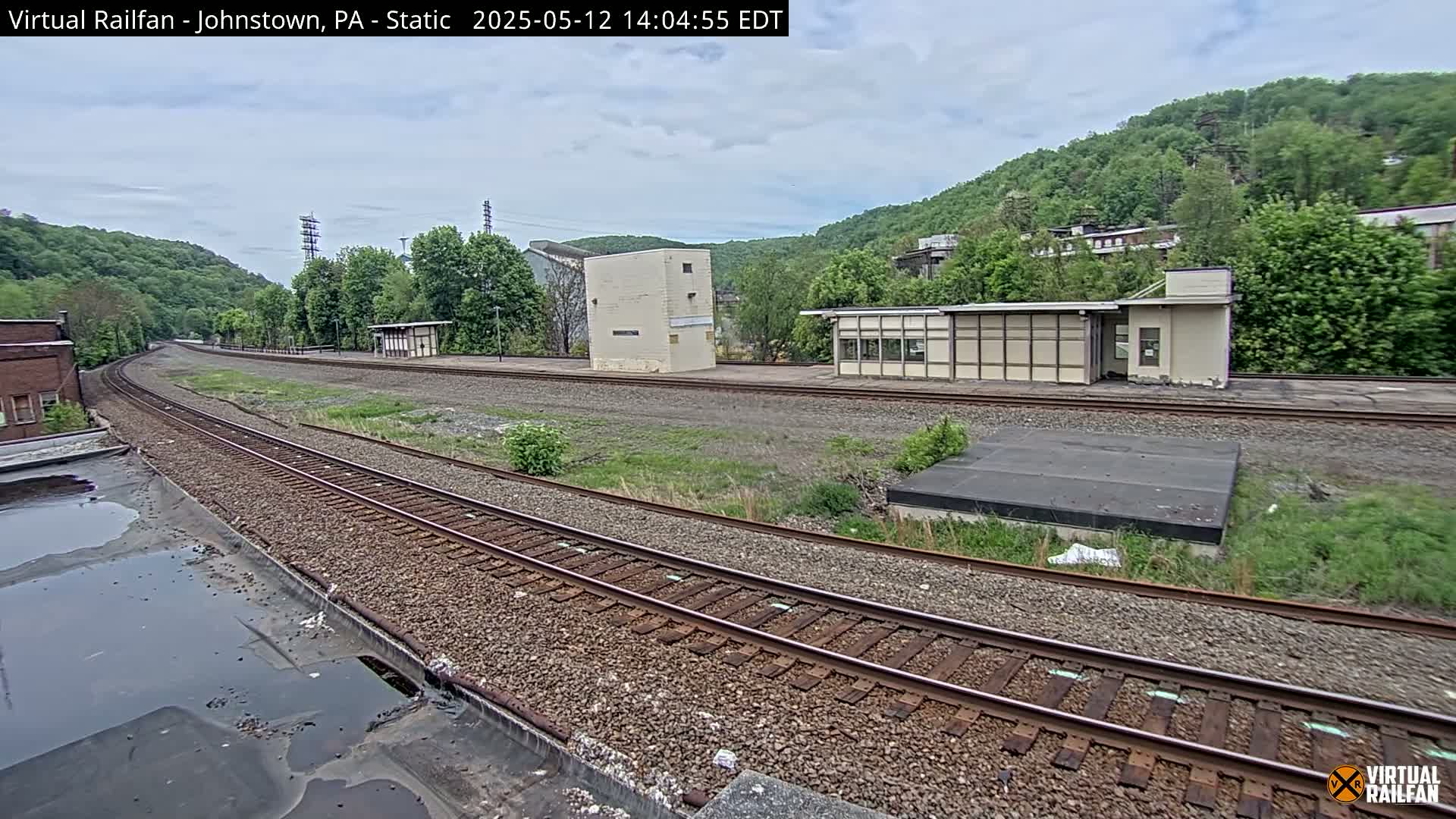 A curving railway track stretches into the distance past several small buildings, nestled against a hillside covered in lush green trees under a partly cloudy sky.