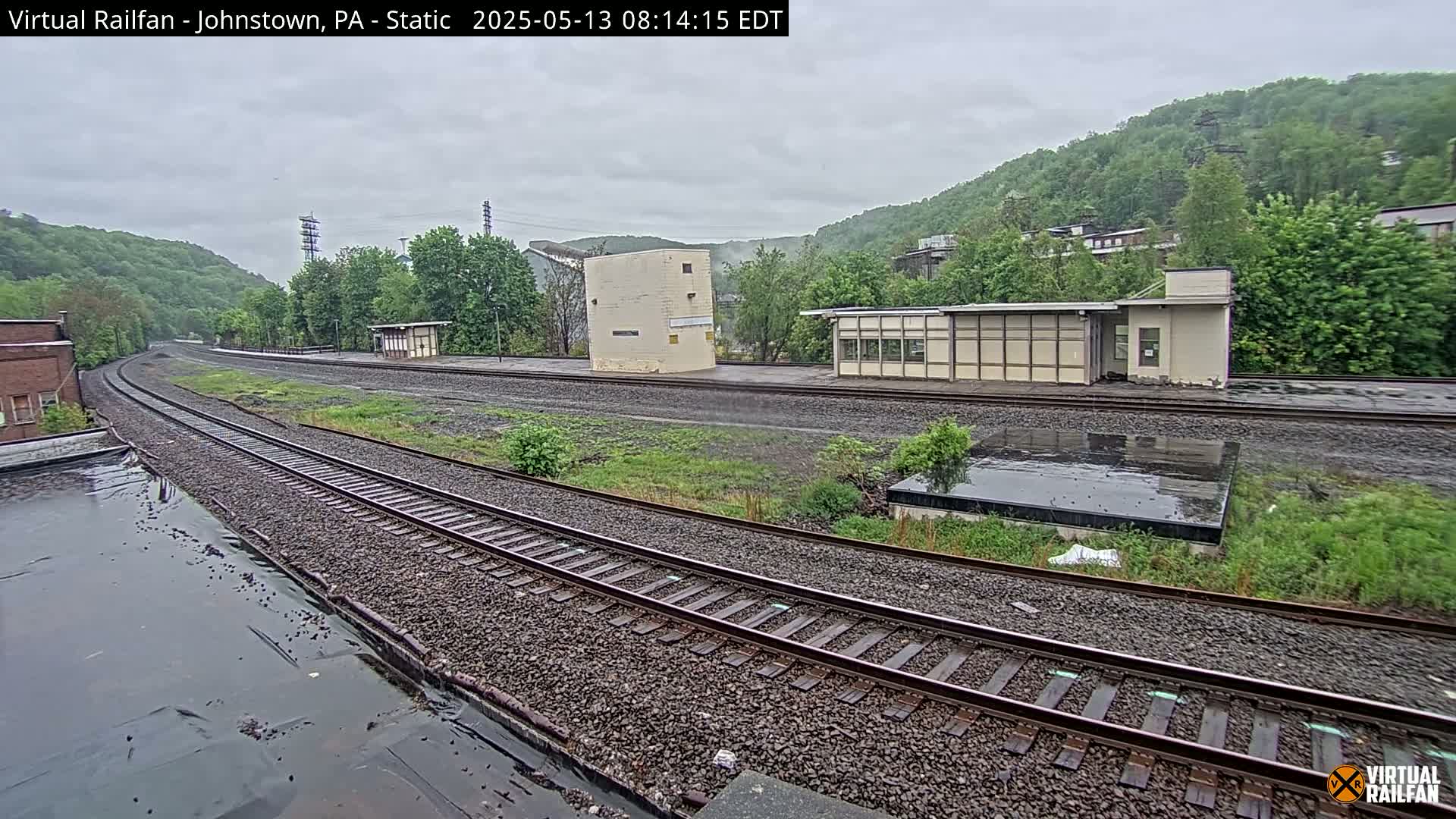 A rain-soaked train yard with multiple tracks curves gently around a small, low building and a larger, boxy structure, all set against a backdrop of lush green hills.